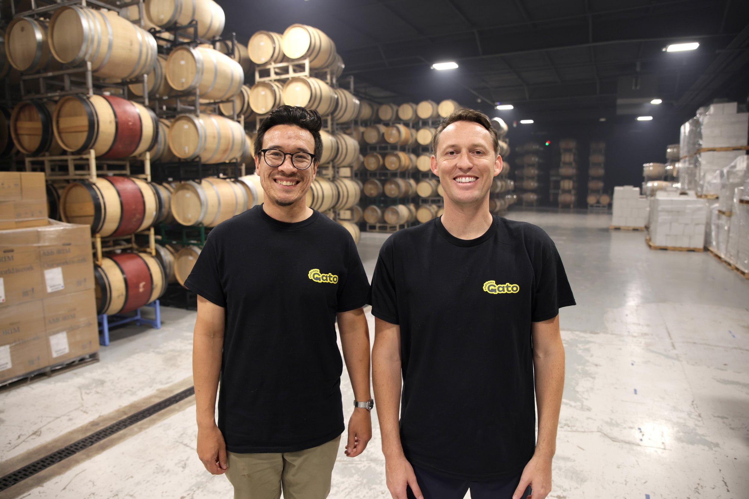 Two men standing inside a warehouse with shelves of wine barrels and stacked boxes in the background, both smiling and wearing black t-shirts.