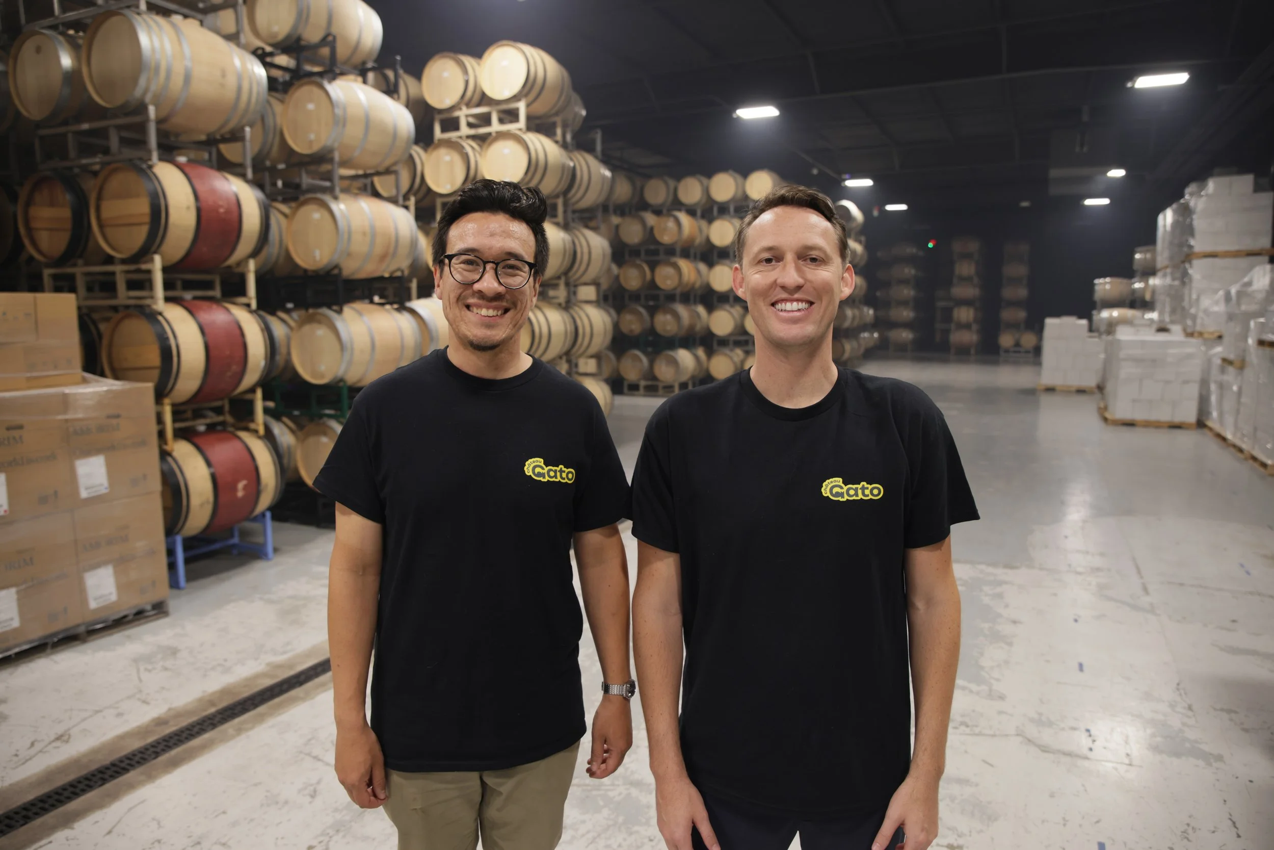 Two men smiling and standing in a warehouse with stacks of barrels and boxes in the background. Both are wearing black t-shirts with a yellow logo.