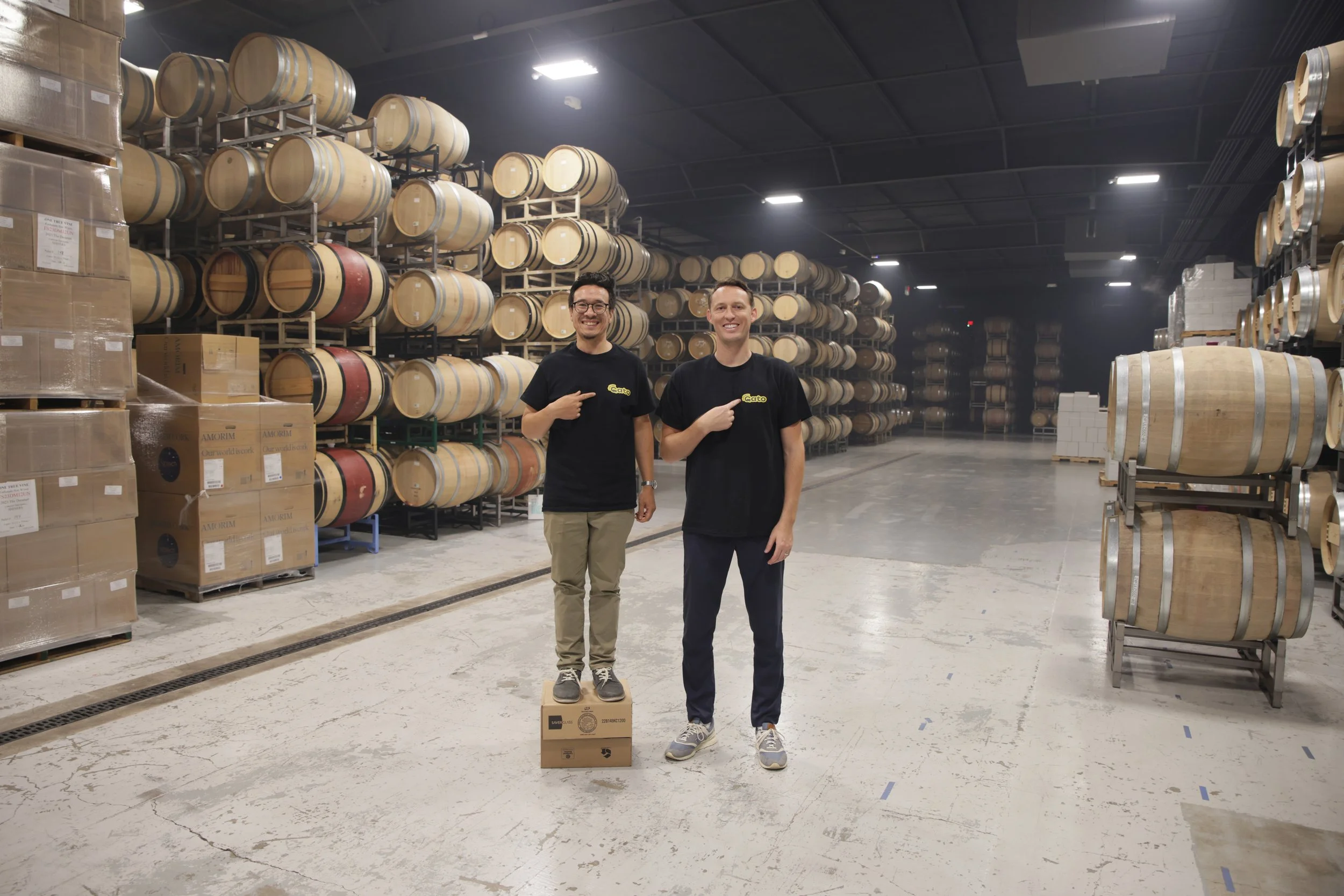 Two men standing in a warehouse filled with stacked wine barrels and boxes, smiling and pointing at each other.