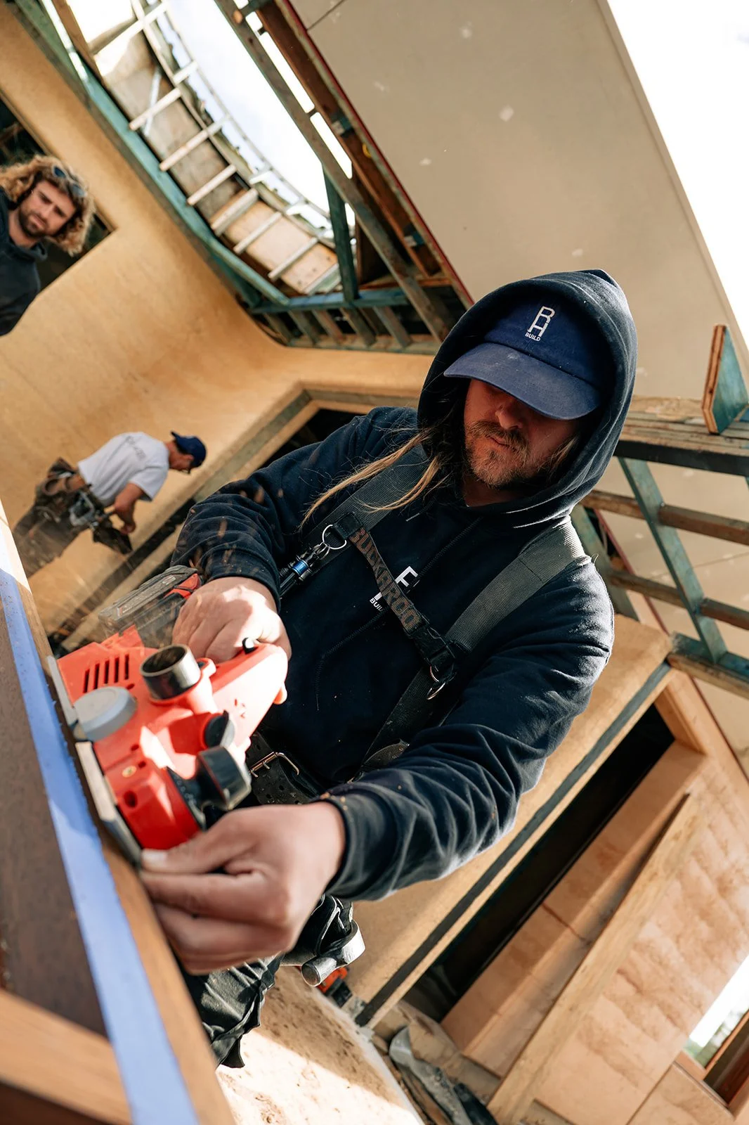 A man wearing a black hoodie and cap using a power tool on a wooden frame inside a building under construction. Two other workers are visible in the background.