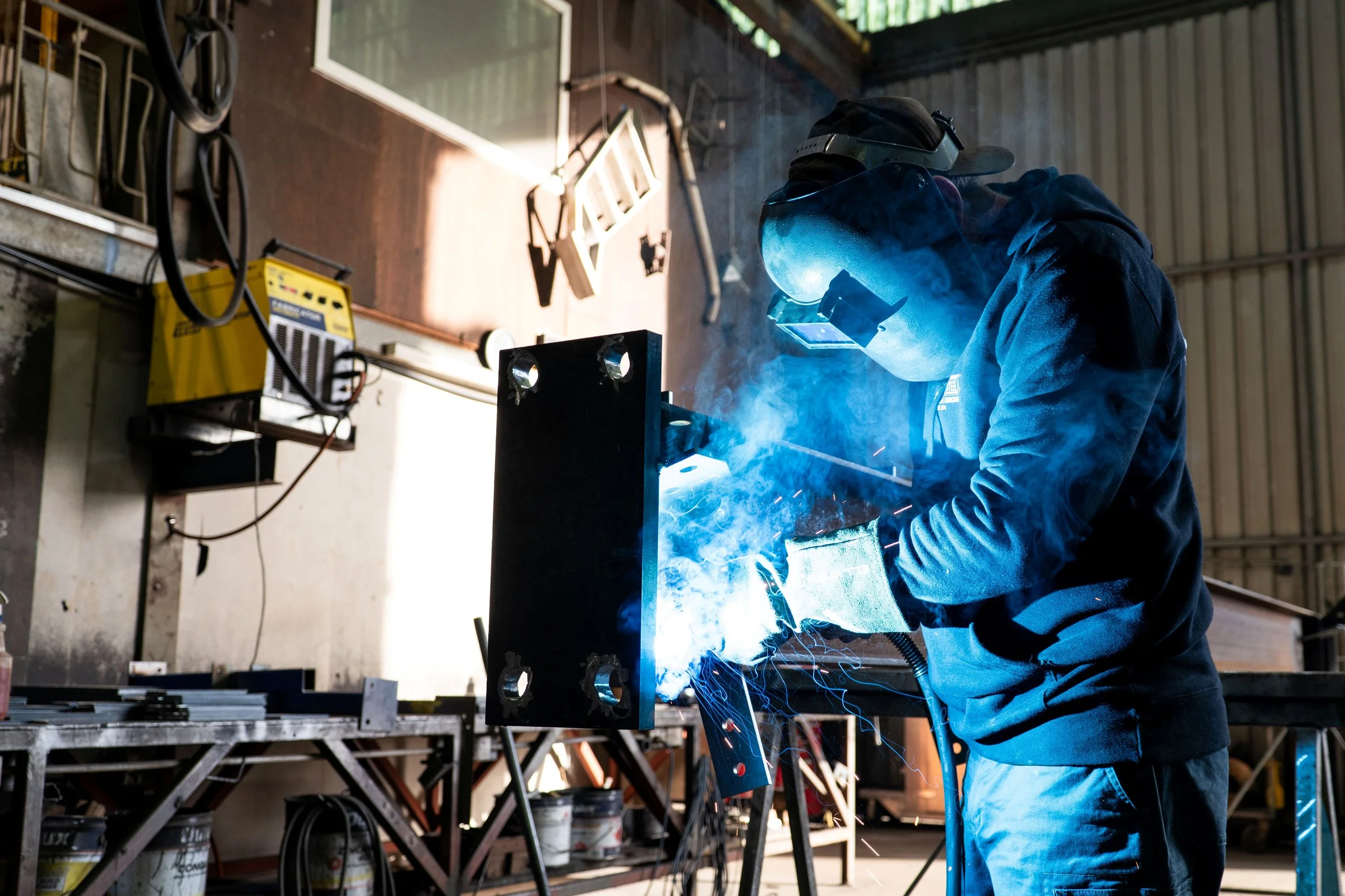 A person welding metal inside a workshop, wearing protective gear including a welding helmet and gloves, with sparks and blue light emitted from the welding process.