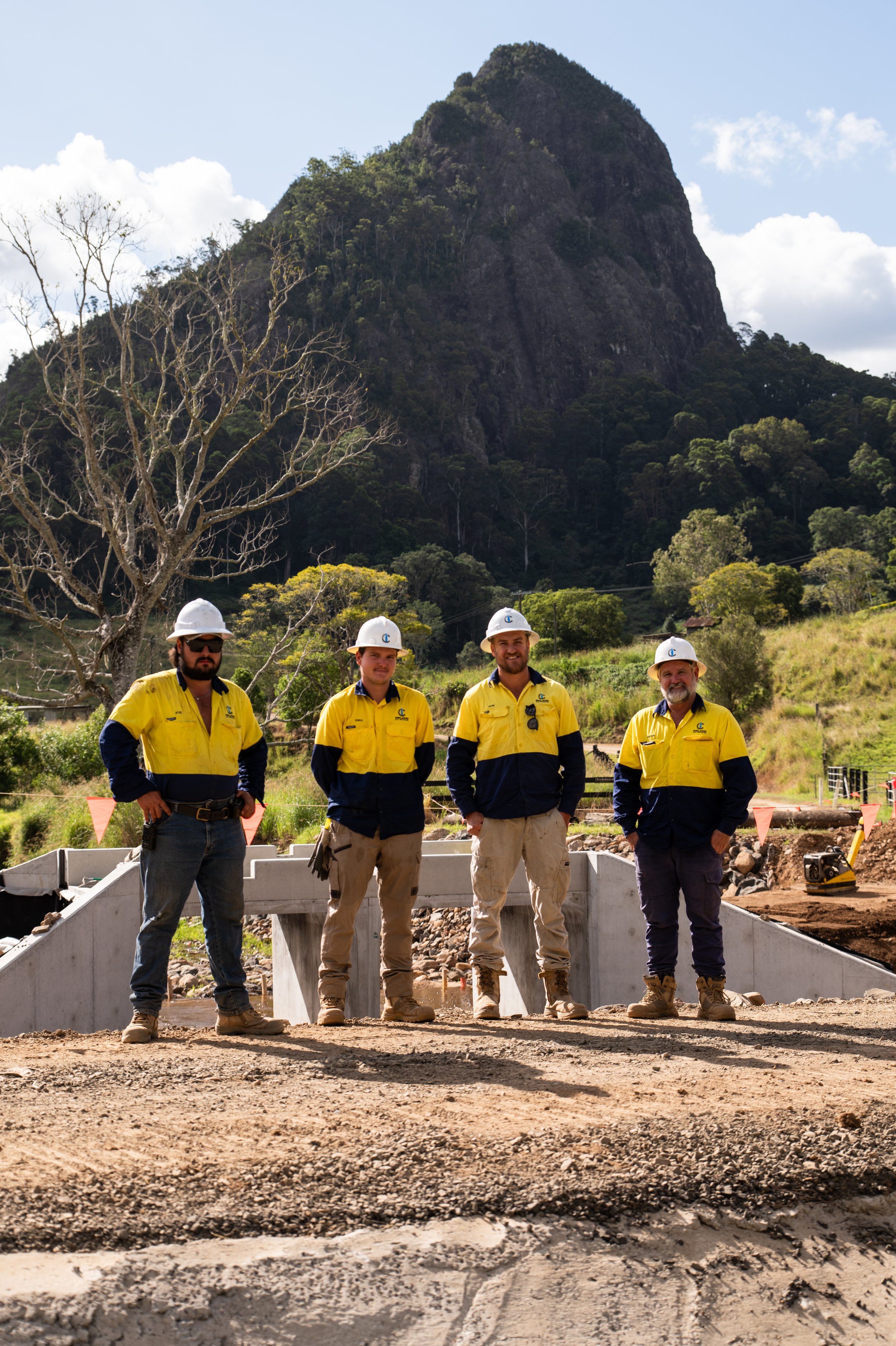 Four construction workers wearing yellow and navy uniforms and white helmets stand in front of a construction site with a mountain in the background. There is a leafless tree and construction equipment around.