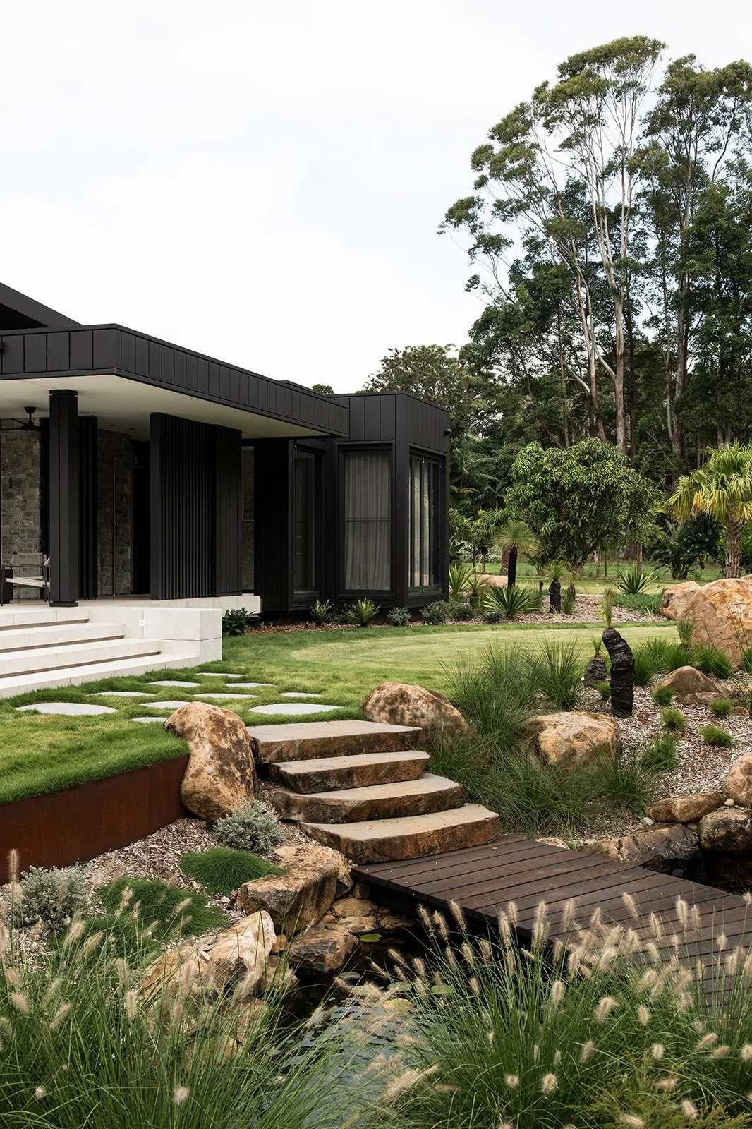 Modern house with black exterior and large windows, surrounded by a landscaped garden with rocks, grass, small plants, and a small pond with a wooden bridge.