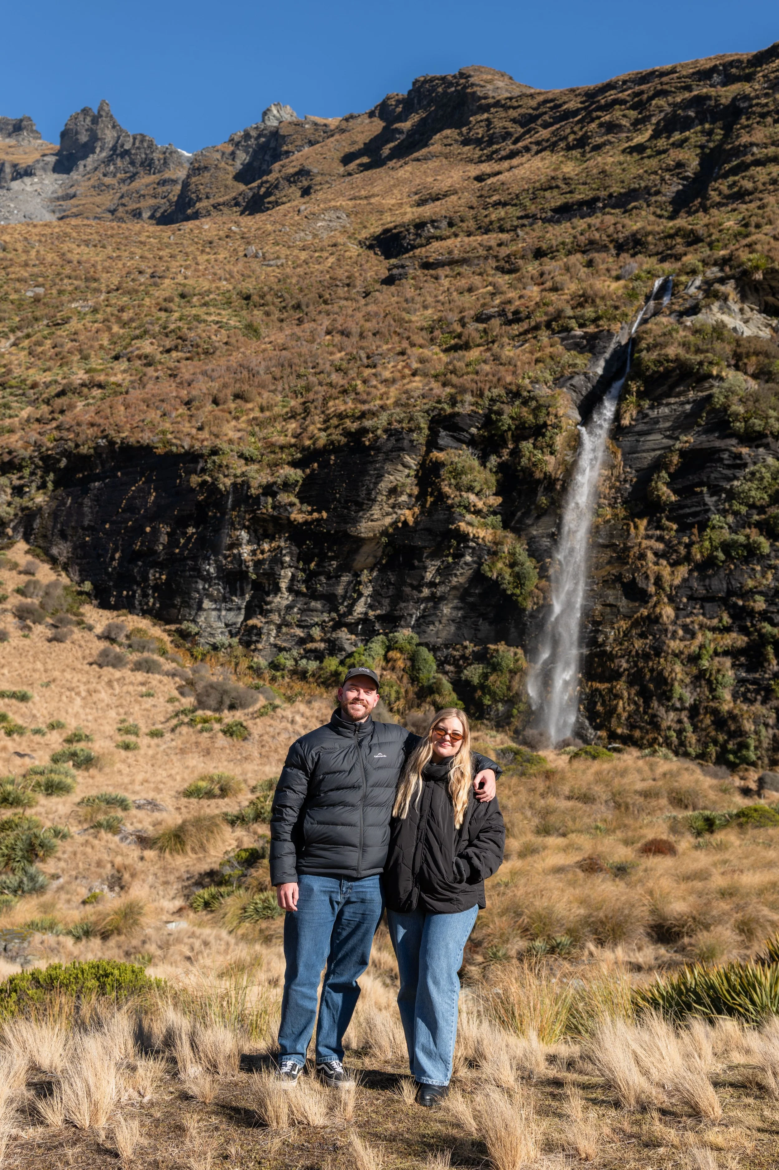 A couple standing outdoors in front of a mountainous landscape with a waterfall. The man is wearing a black jacket and jeans, and the woman is wearing sunglasses, a black jacket, and jeans. They are smiling and have their arms around each other.