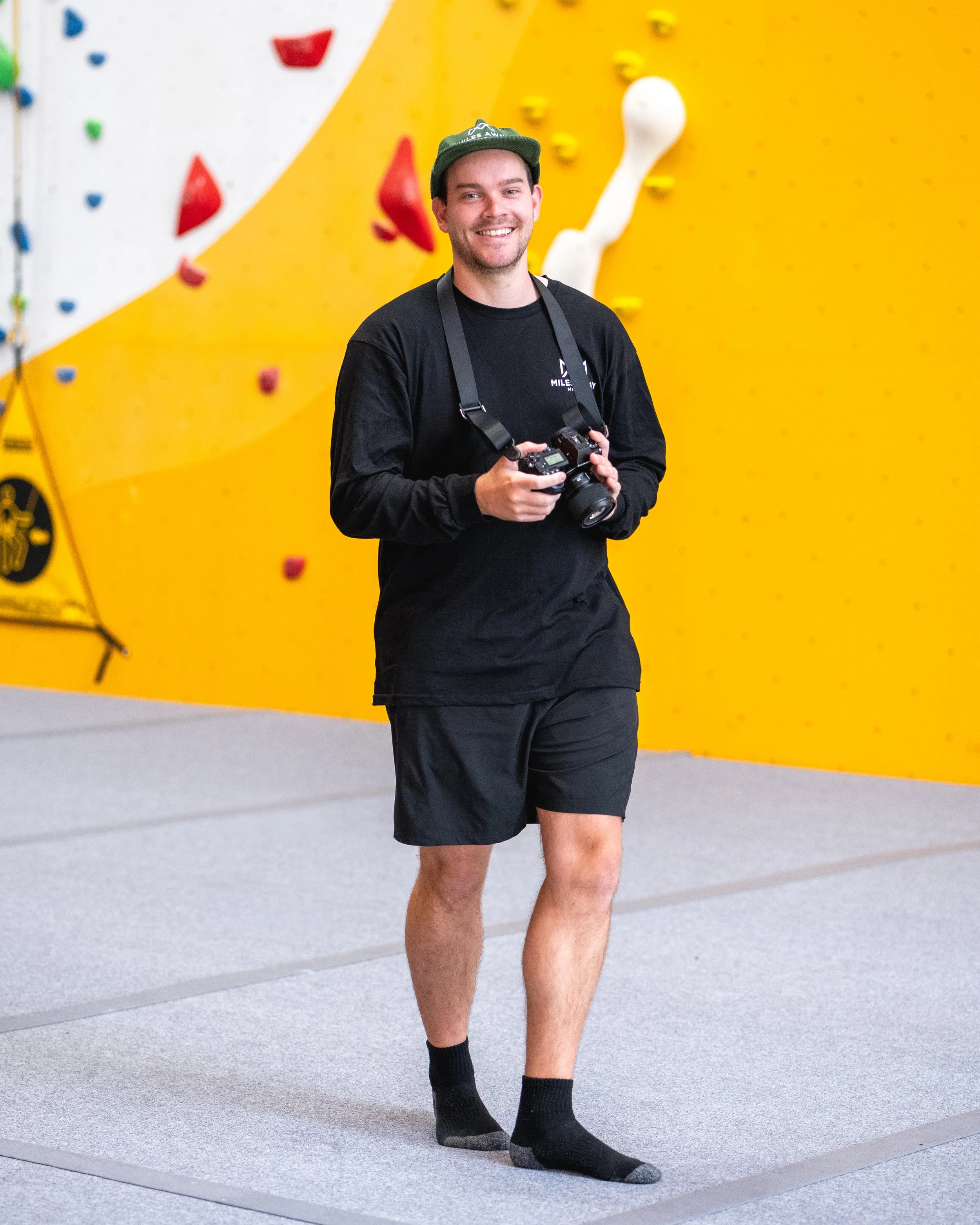 A young man with short brown hair, smiling, holding a camera, wearing a green cap, black long-sleeve shirt, black shorts, and black socks, standing on a gray indoor climbing gym with a yellow climbing wall in the background.