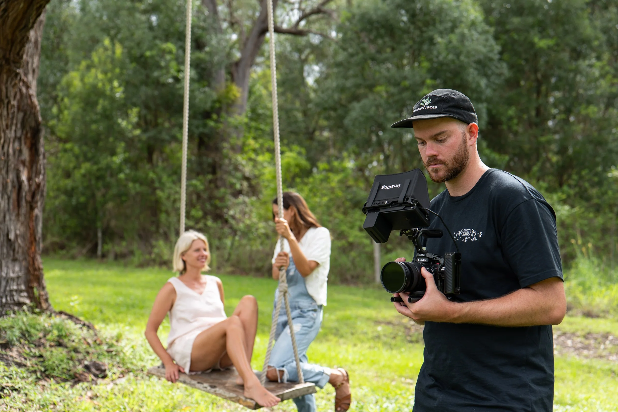 A man with a camera filming a woman sitting on a swing in a park with two other women standing nearby, all surrounded by green trees.