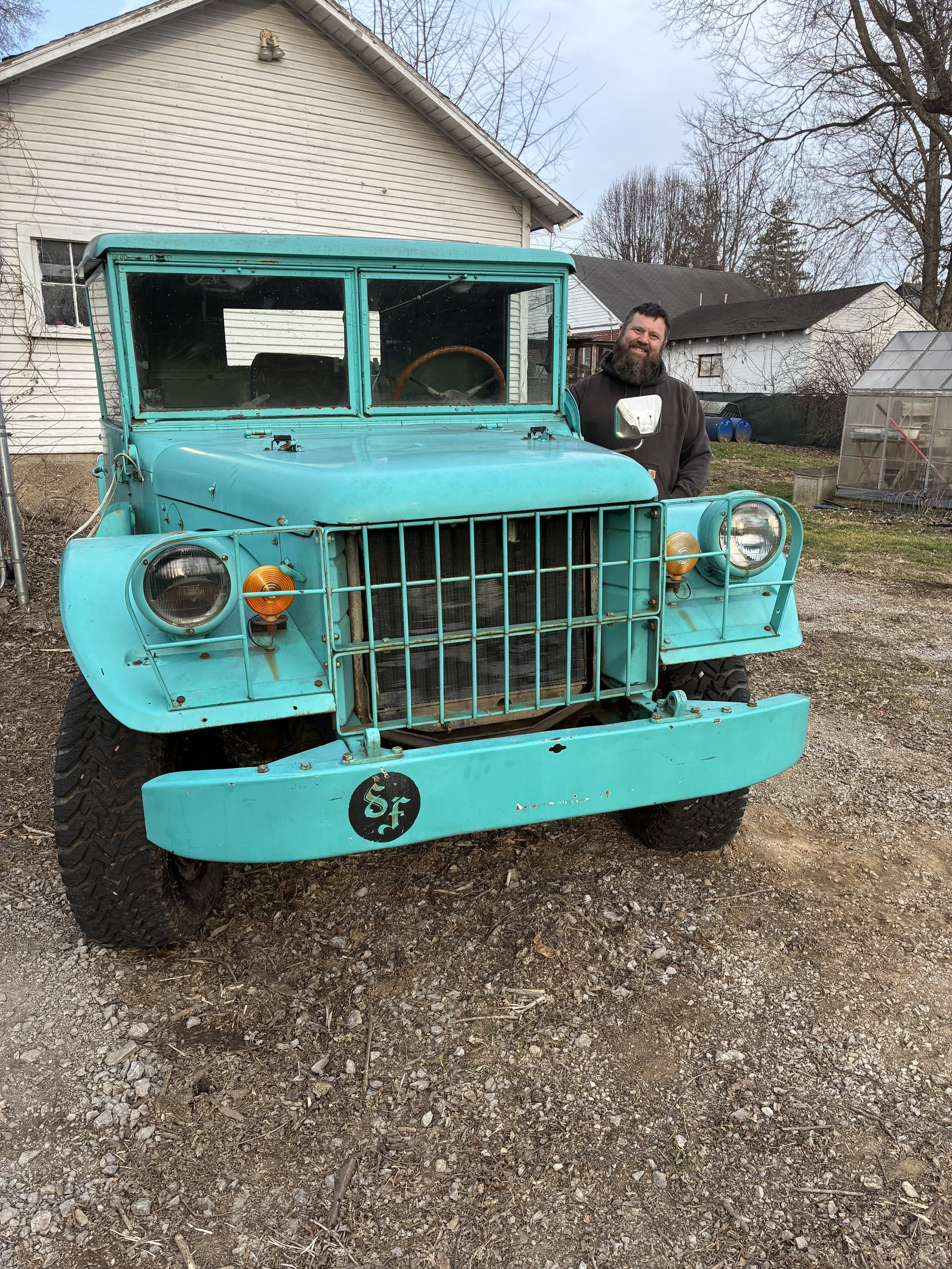 A man with a beard standing next to a vintage turquoise utility vehicle in a backyard with houses, trees, and a greenhouse in the background.