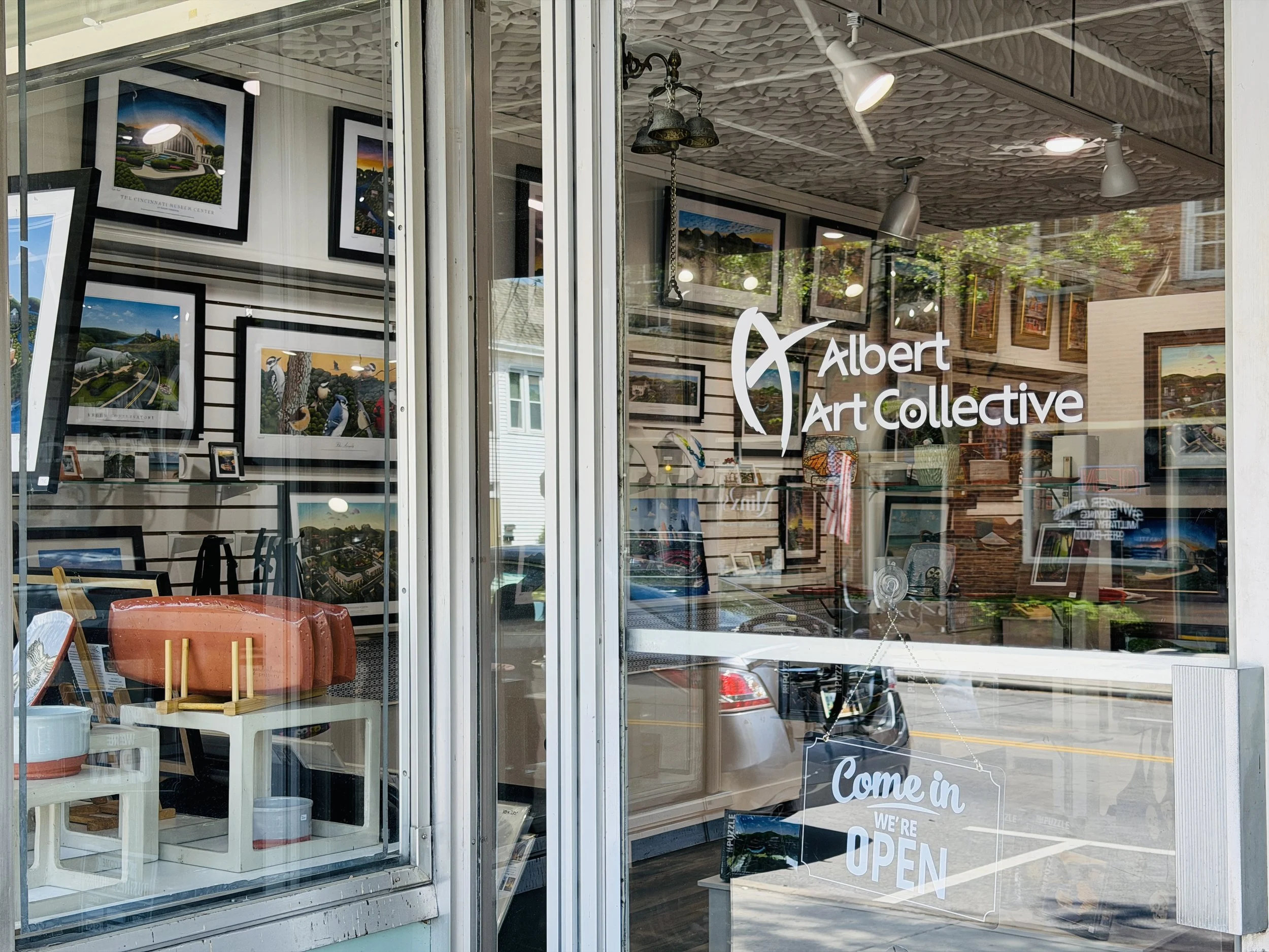 View of an art gallery storefront with artwork displayed inside and a sign on the glass door reading 'Come in, we're open' and 'Albert Art Collective'.