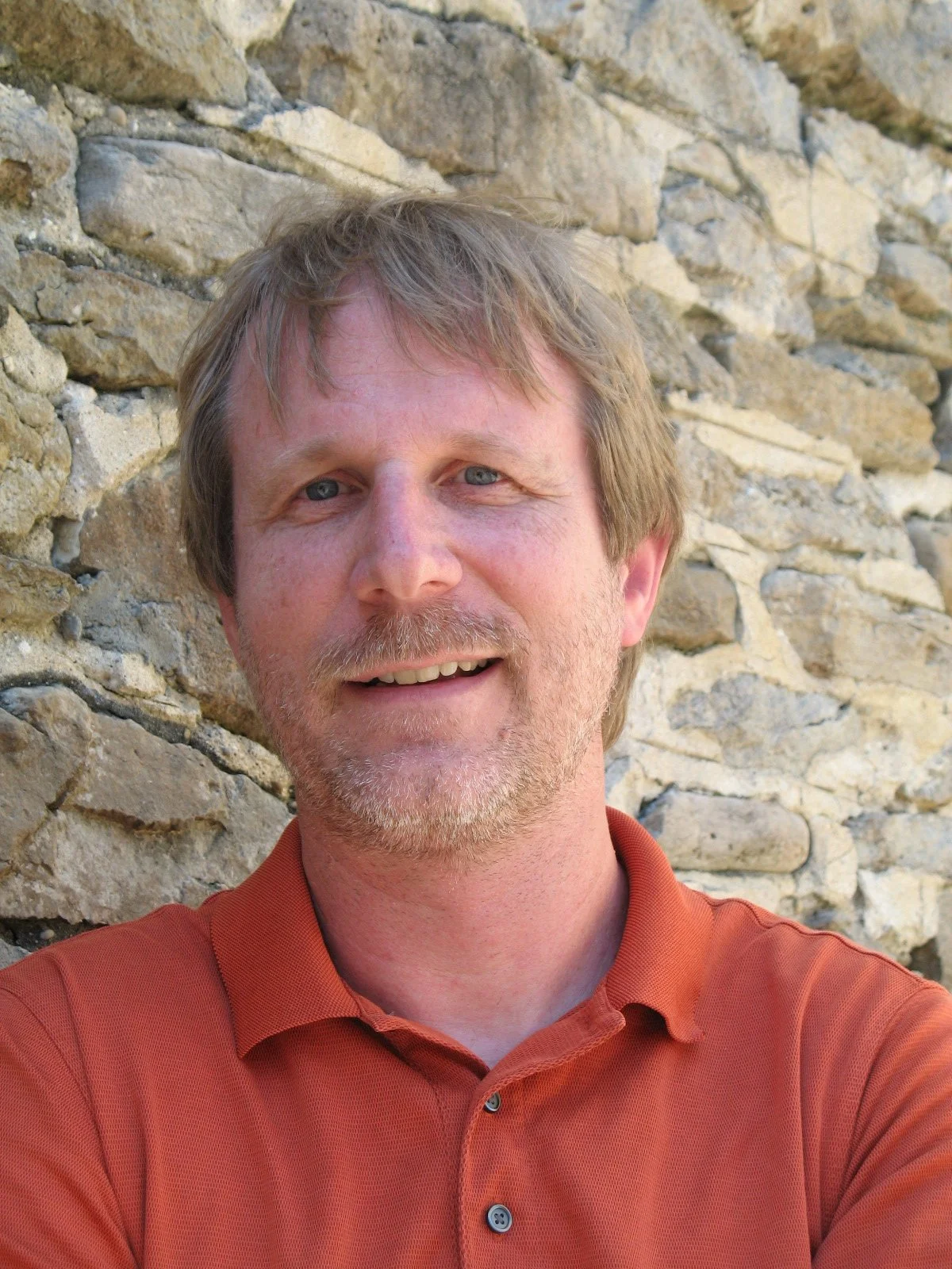 A middle-aged man with light brown hair and a beard, smiling, wearing an orange collared shirt, standing in front of a stone wall.