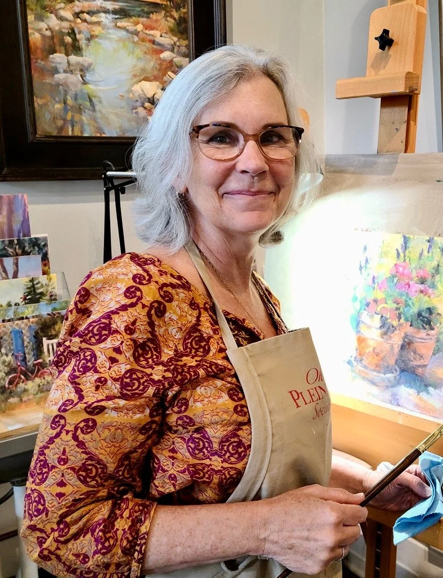 An elderly woman painting outdoors on a street sidewalk. She is wearing a pink cap, glasses, and a sleeveless brown shirt, holding a paintbrush and palette, with her painting setup on an easel. Behind her are cars parked on the street, trees, and buildings with striped awnings.