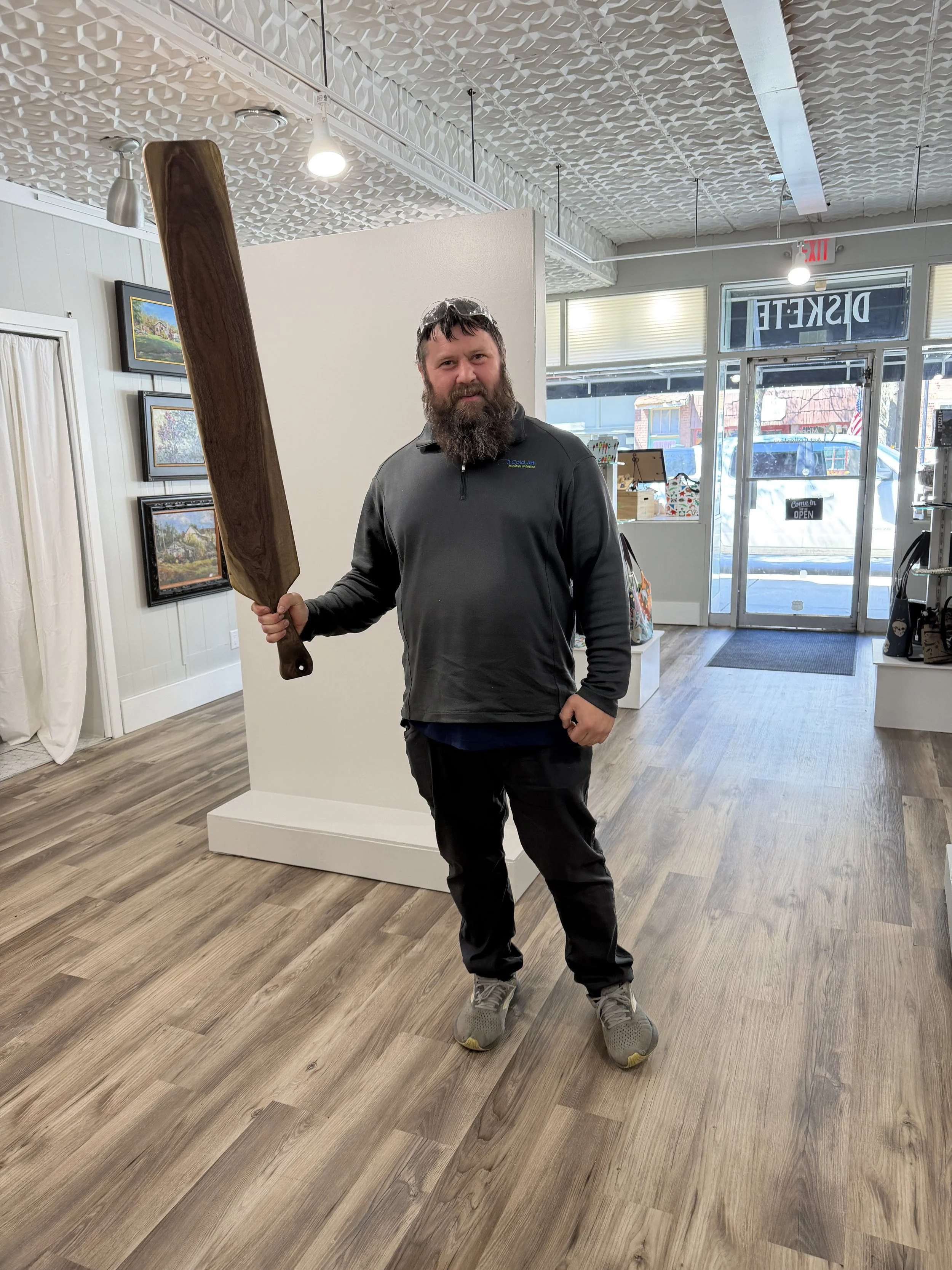 A man with a beard and glasses on his head is standing indoors, holding a large wooden paddle, in front of a display wall. He is wearing a black jacket, black pants, and gray athletic shoes. The setting appears to be a shop or gallery with artwork on the walls, a glass door with a sign, and a wooden floor.