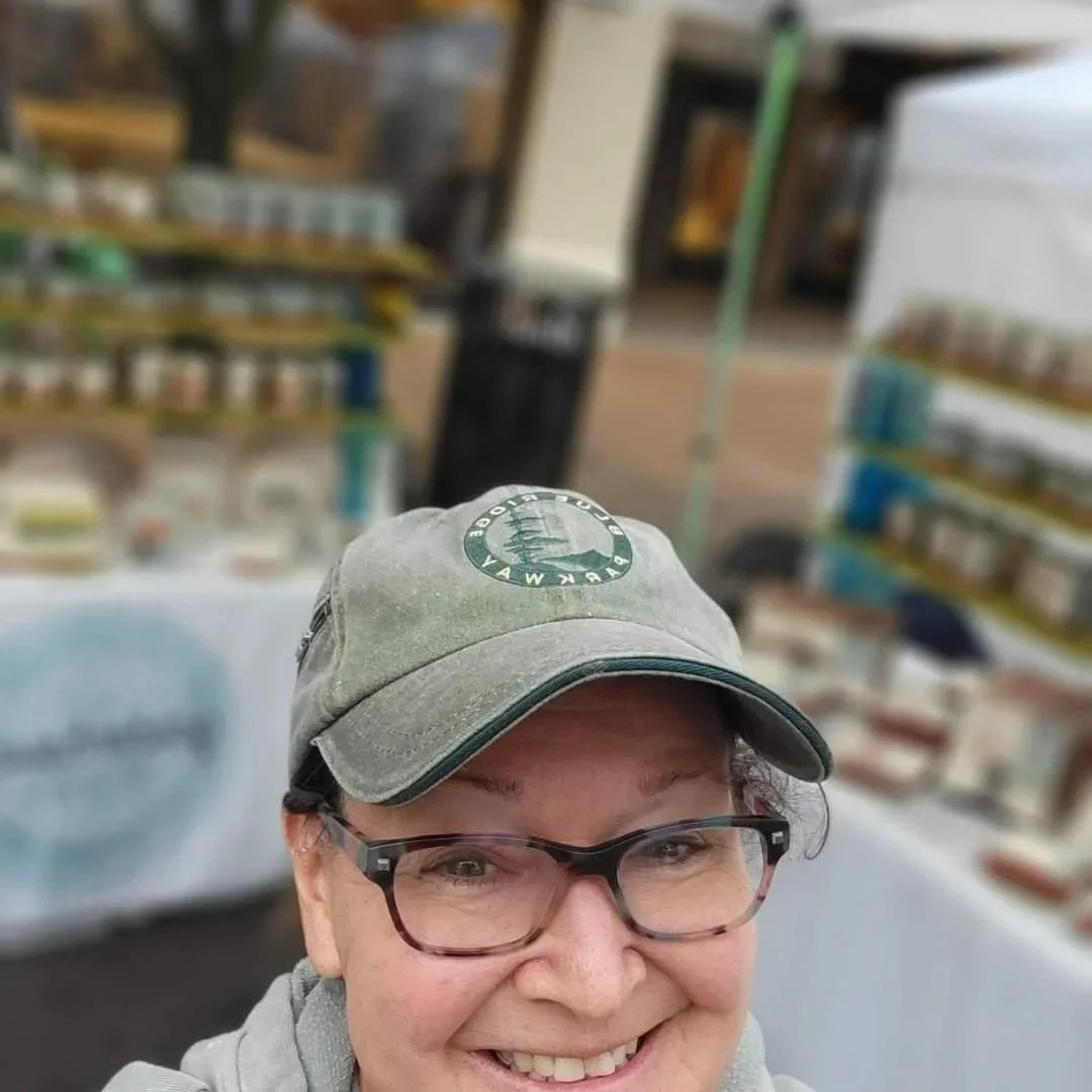 A woman taking a selfie outdoors near a store, wearing glasses and a gray cap with a logo, smiling at the camera.