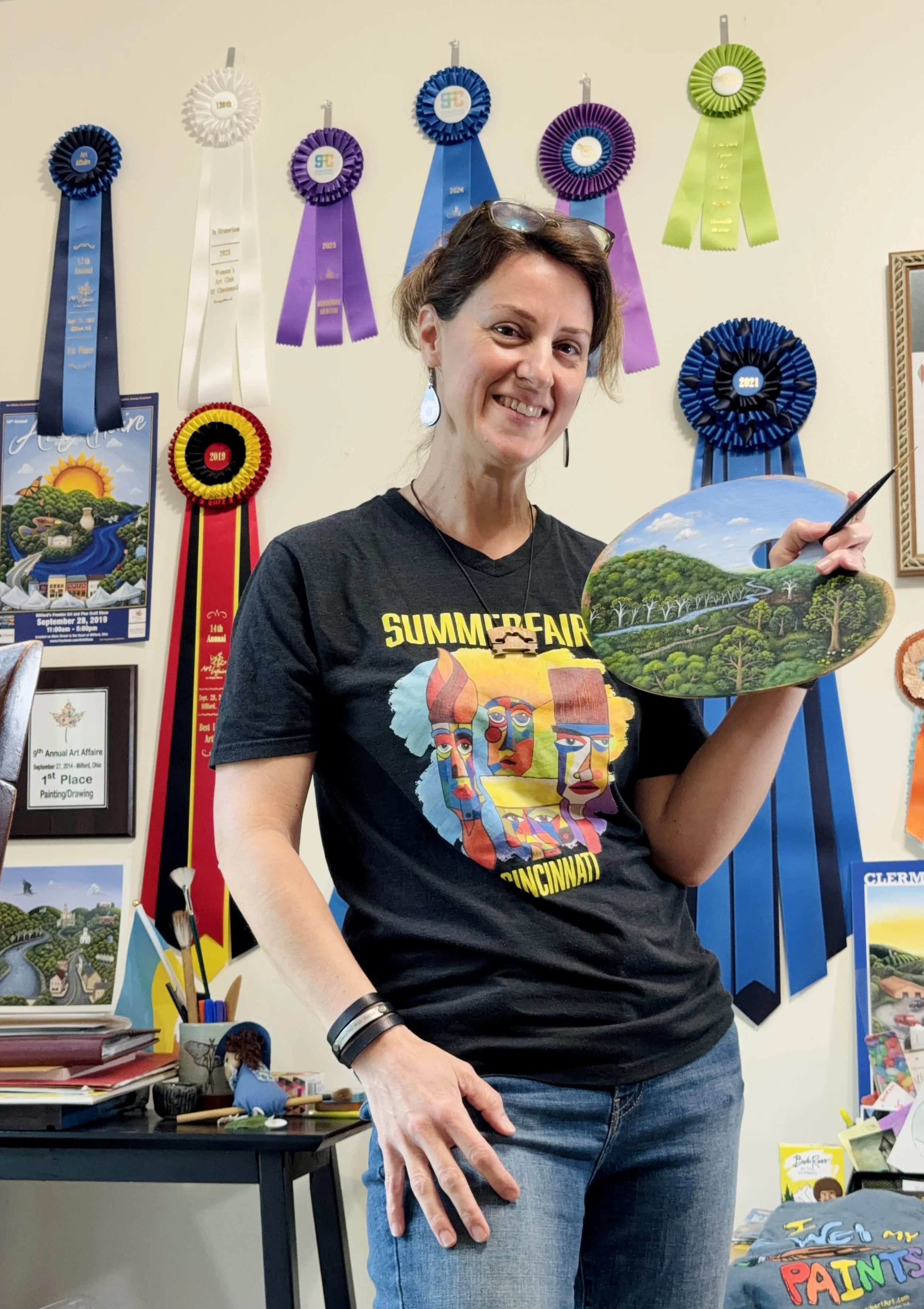 Woman in a black t-shirt with colorful art designs holds a painting of a landscape with trees, sky, and river. She stands in front of a wall decorated with award ribbons and artwork.