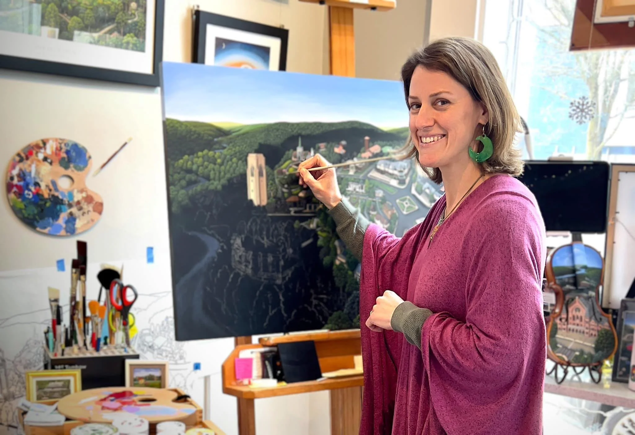 A woman painting on a large outdoor mural, smiling and wearing a pink cardigan, in an art studio filled with paintings, art supplies, and framed artwork.