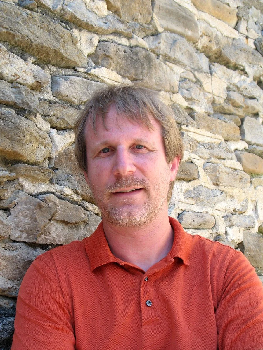 A middle-aged man with light brown hair, blue eyes, and a beard, wearing an orange polo shirt, standing outdoors in front of a stone wall.
