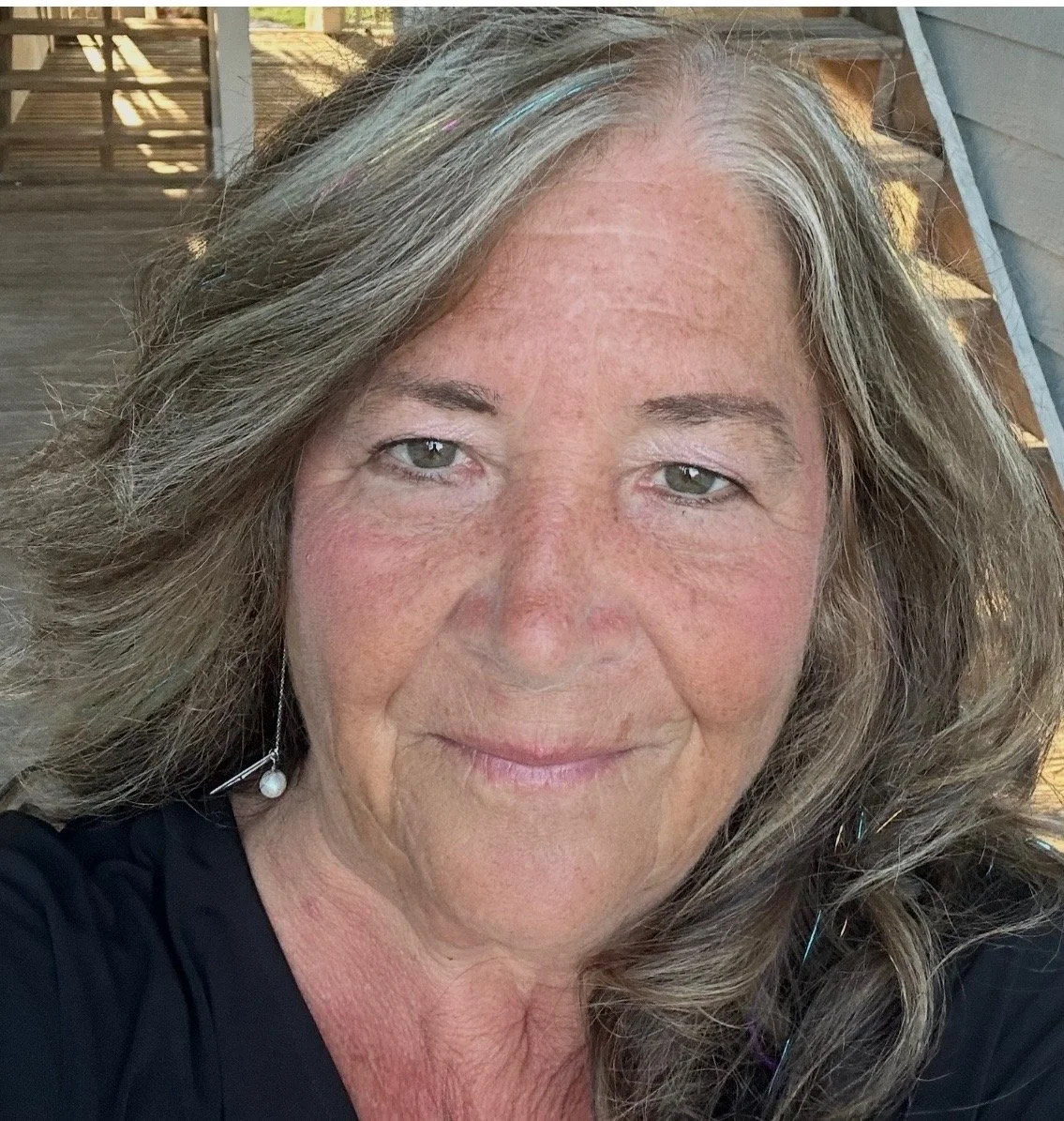 Close-up of a middle-aged woman with wavy brown hair with gray streaks, light skin, and wearing a black top and pearl earring, outdoors with a wooden deck and stairs in the background.