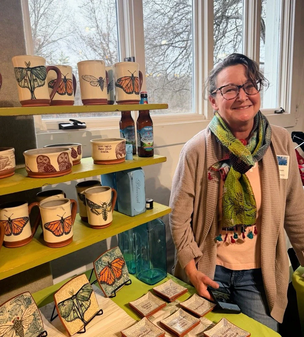A woman with short gray hair, glasses, wearing a multicolored scarf and beige jacket, standing at a display table with handmade ceramics and small plaques, in front of windows showing trees outside.