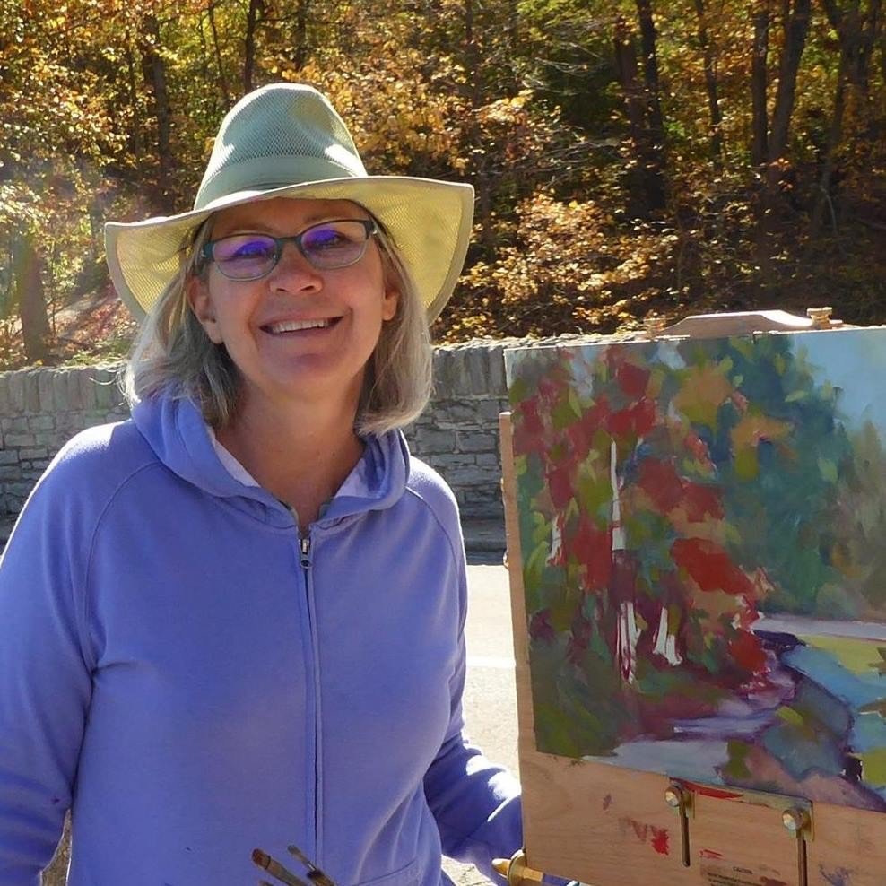 A woman with blonde hair, wearing a wide-brimmed hat, glasses, and a lavender hoodie, smiling while standing outdoors next to an easel with a colorful landscape painting, in front of a stone wall and trees with autumn leaves.