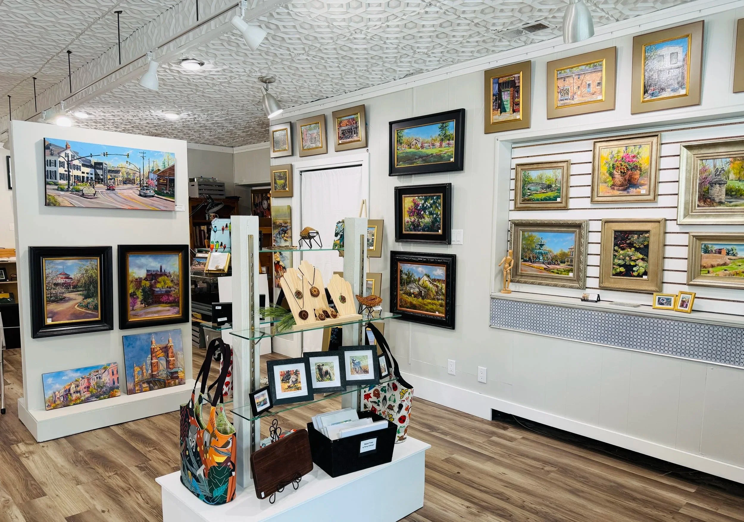 An art gallery display with framed landscape paintings on the walls and smaller artworks on tables and shelves, with a wooden floor and ceiling lighting.