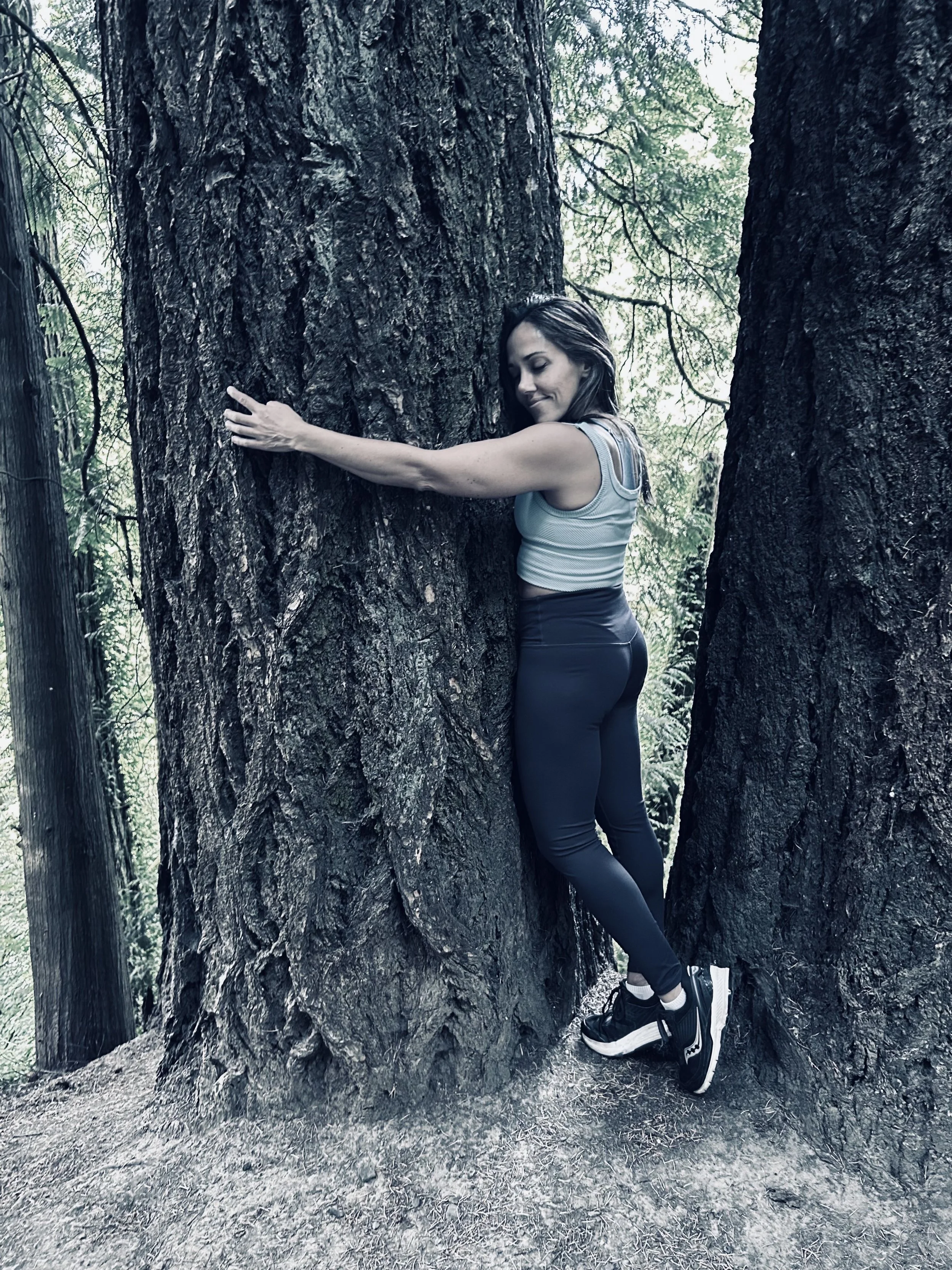 Woman hugging a large tree in a forest, wearing athletic clothing and sneakers.