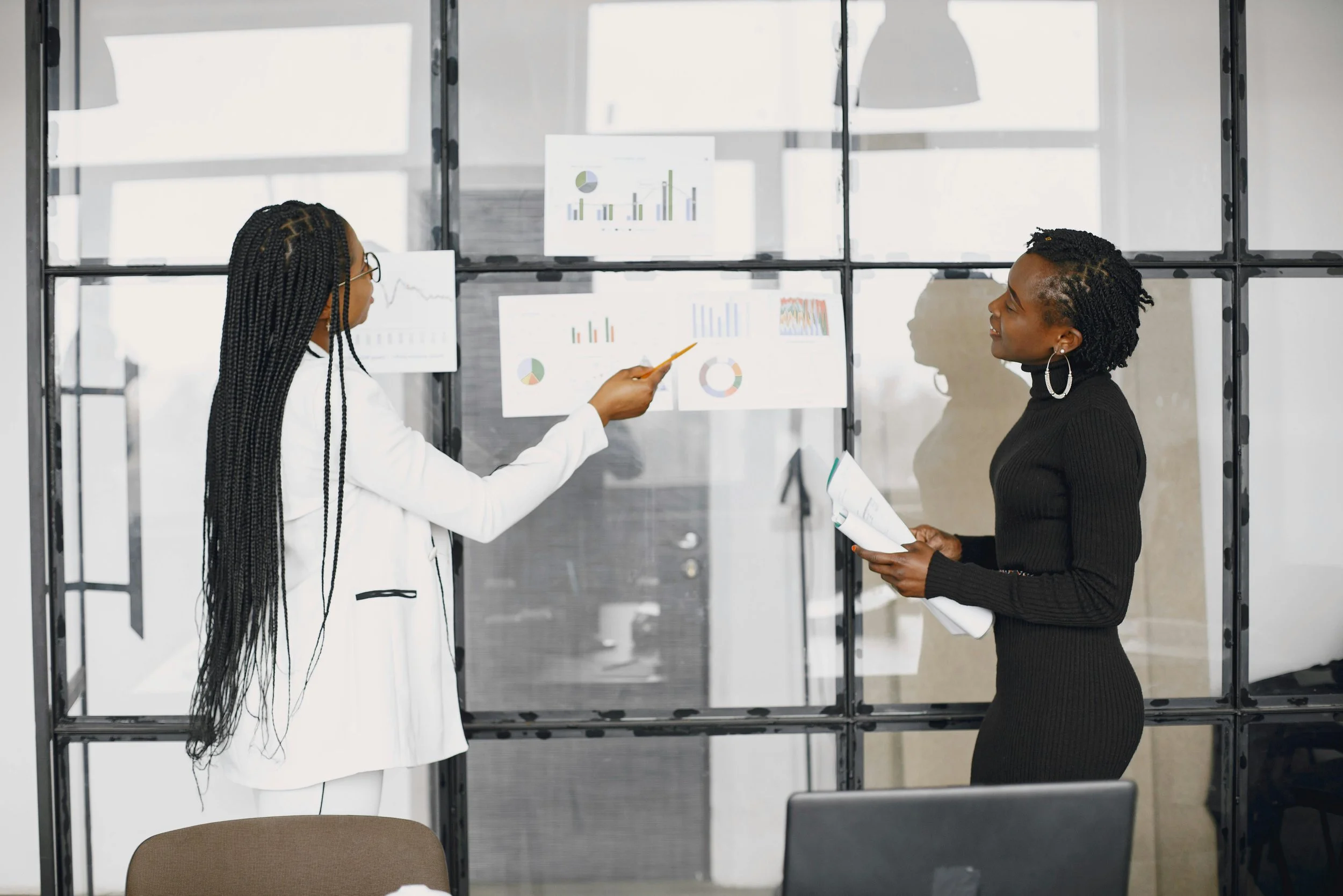 Two women having a discussion in front of data charts displayed on a glass wall in a modern office.