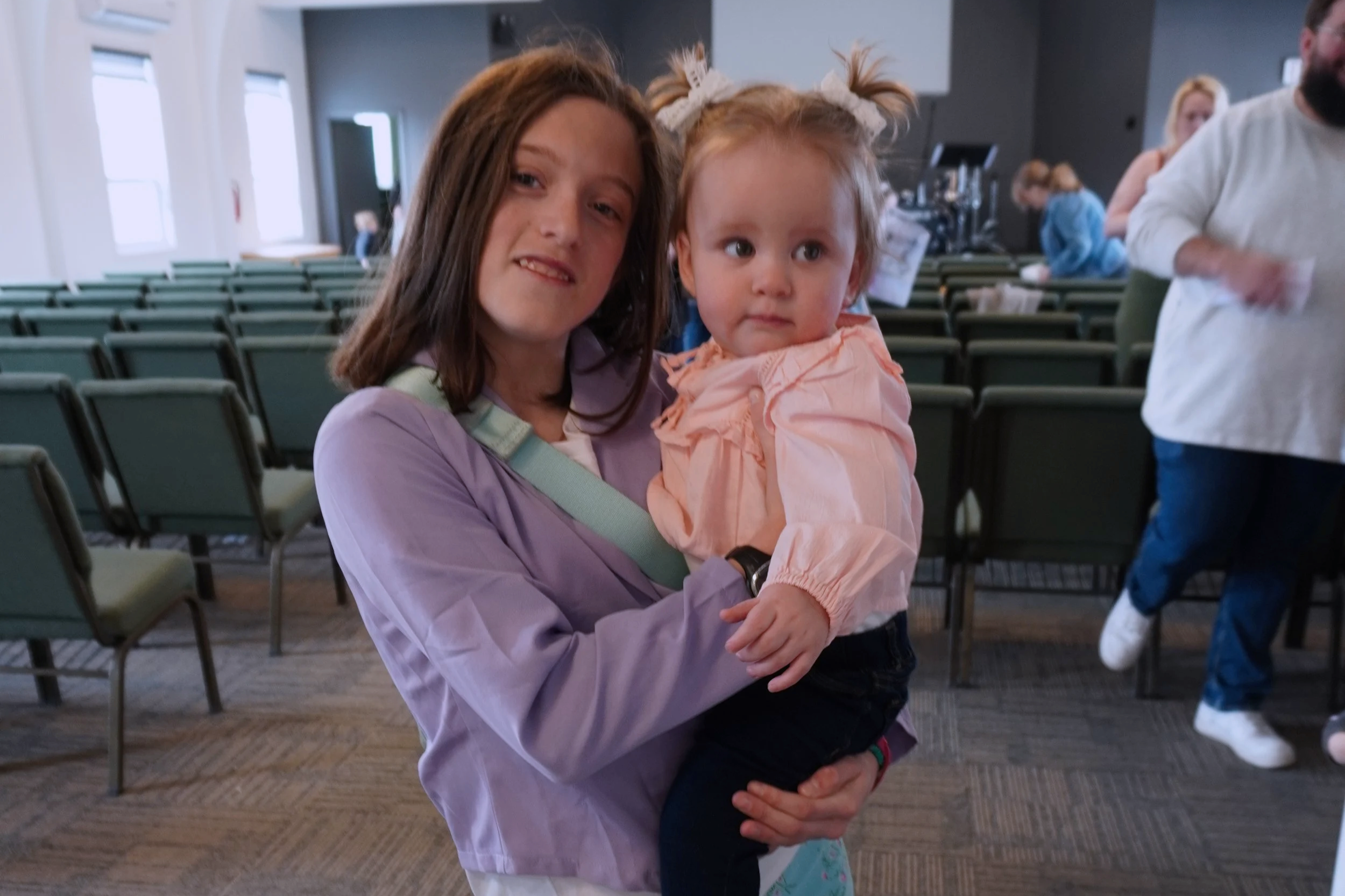 A woman with brown hair holding a young girl with red hair and pigtails in an indoor setting with empty chairs and other people in the background.