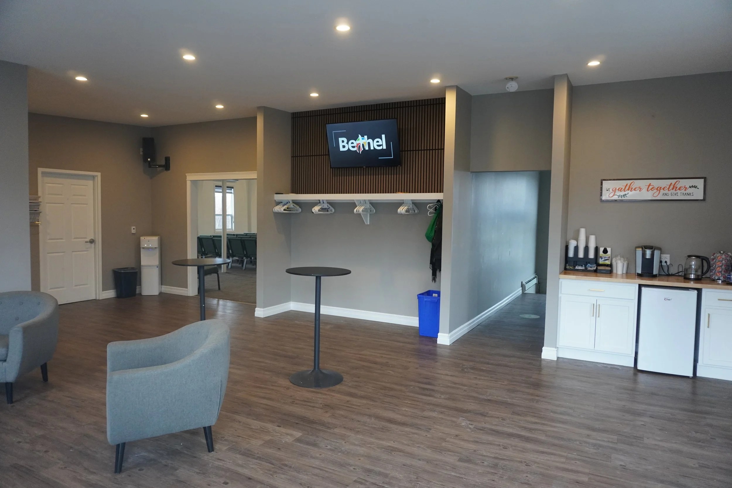 Indoor communal space with chairs, tables, a kitchen area with cups and a coffee maker, a wall-mounted TV displaying 'Bethel' logo, and decorative signs.