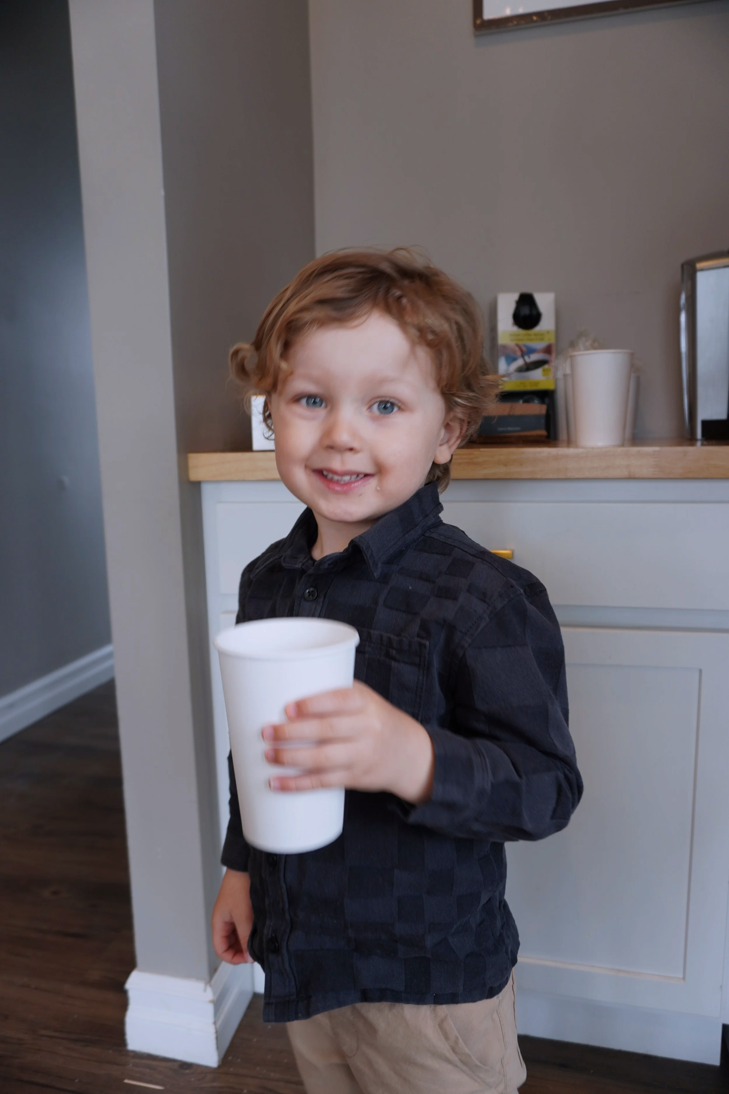 Young boy with curly red hair and blue eyes smiling, holding a white cup, in a modern kitchen with wood and white cabinetry.