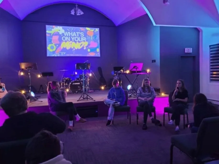 A panel of five young women sitting on chairs in front of a small audience, engaged in a discussion on a stage in a dimly lit room with blue and purple lighting, musical instruments, and a screen displaying the question "What's on your mind?".