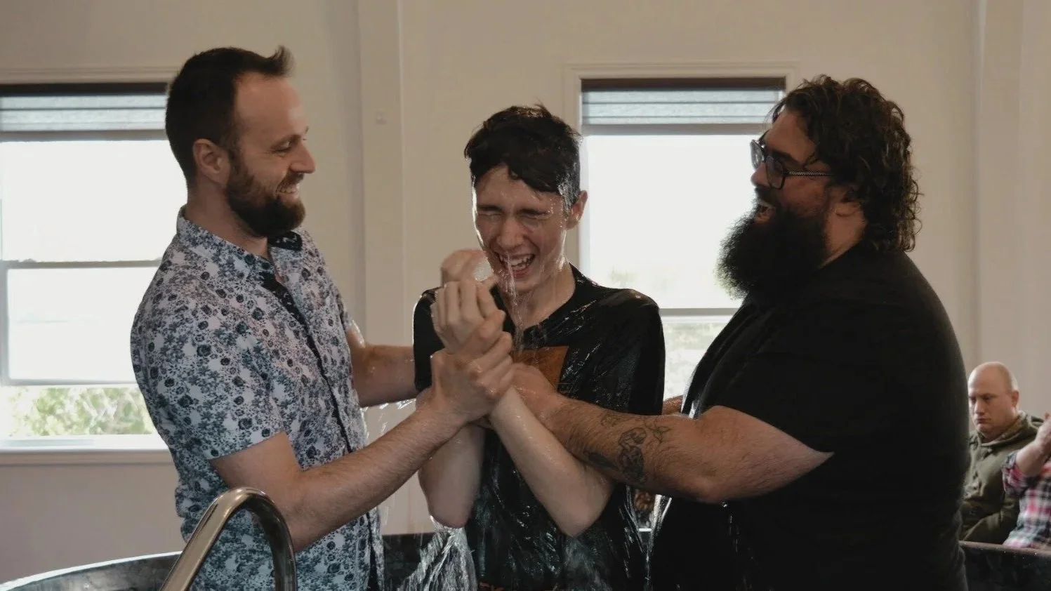 Three men smiling and celebrating during a baptism ceremony indoors, water splashing on them