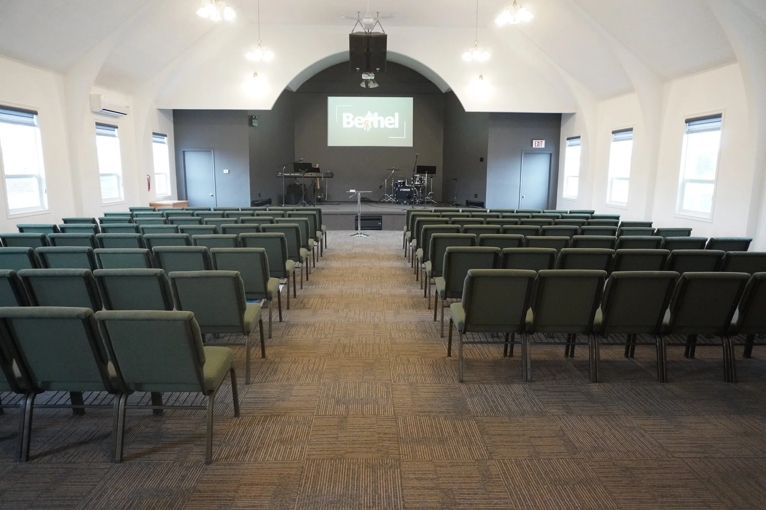 Empty auditorium or conference room with rows of chairs facing a stage with musical instruments and a large screen displaying the word 'Behel'.