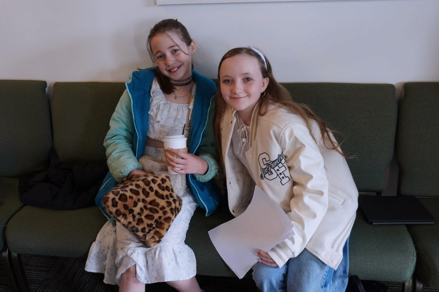 Two young girls sitting on a green bench, smiling at the camera. One girl is holding a disposable coffee cup and a leopard print blanket. The girl on the right is wearing a white jacket with a letter 'F' and the word 'Southampton' on it, and is holding a sheet of paper. The background shows a plain wall and part of a black bag on the left side.