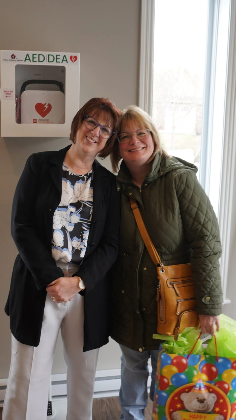 Two women smiling and standing close together indoors, with a window behind them. One woman is wearing glasses, a black jacket, a floral blouse, and white pants. The other woman is wearing glasses, an olive green quilted jacket, and is holding colorful gift bags with a birthday theme.