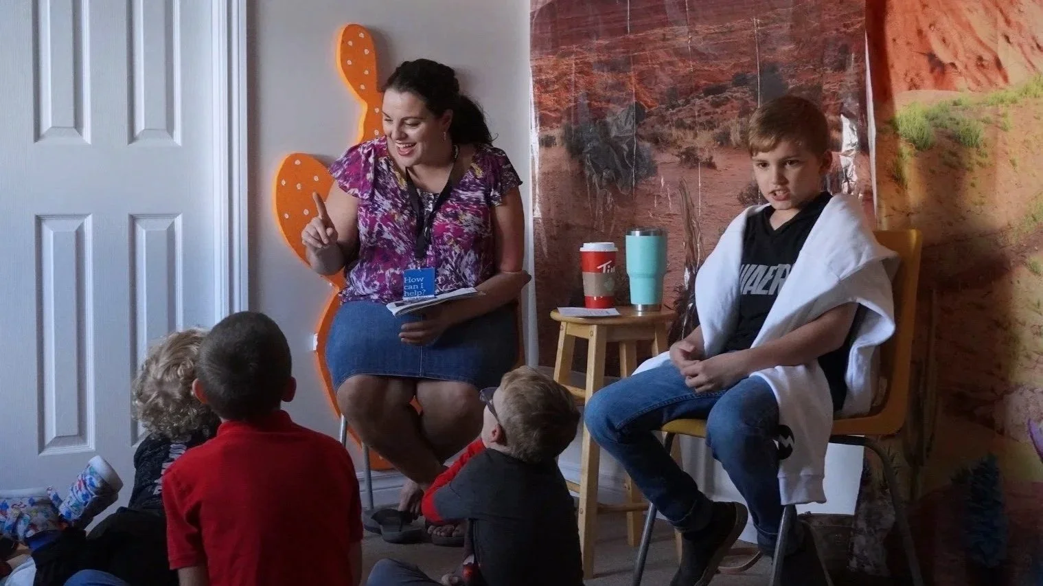 A woman reading a book to children in a classroom or library setting, with some children sitting on the floor and one boy sitting on a chair. The woman is smiling and holding a book, and there are cups on a nearby small table.