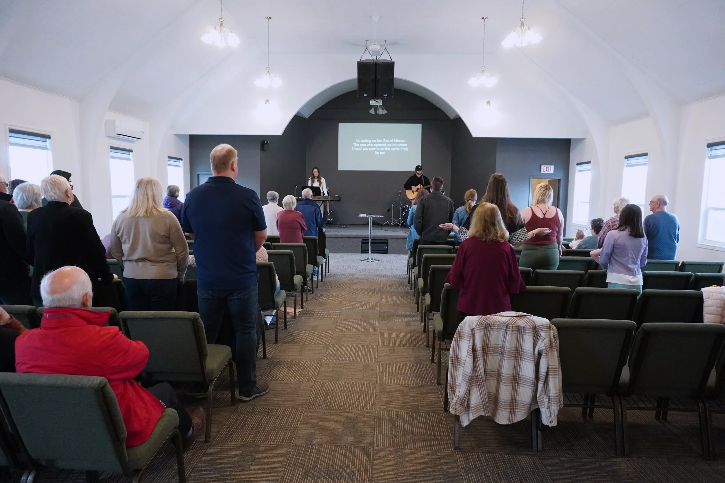 People gathered in a church or auditorium listening to a live musical performance on stage, with lyrics displayed on a screen.