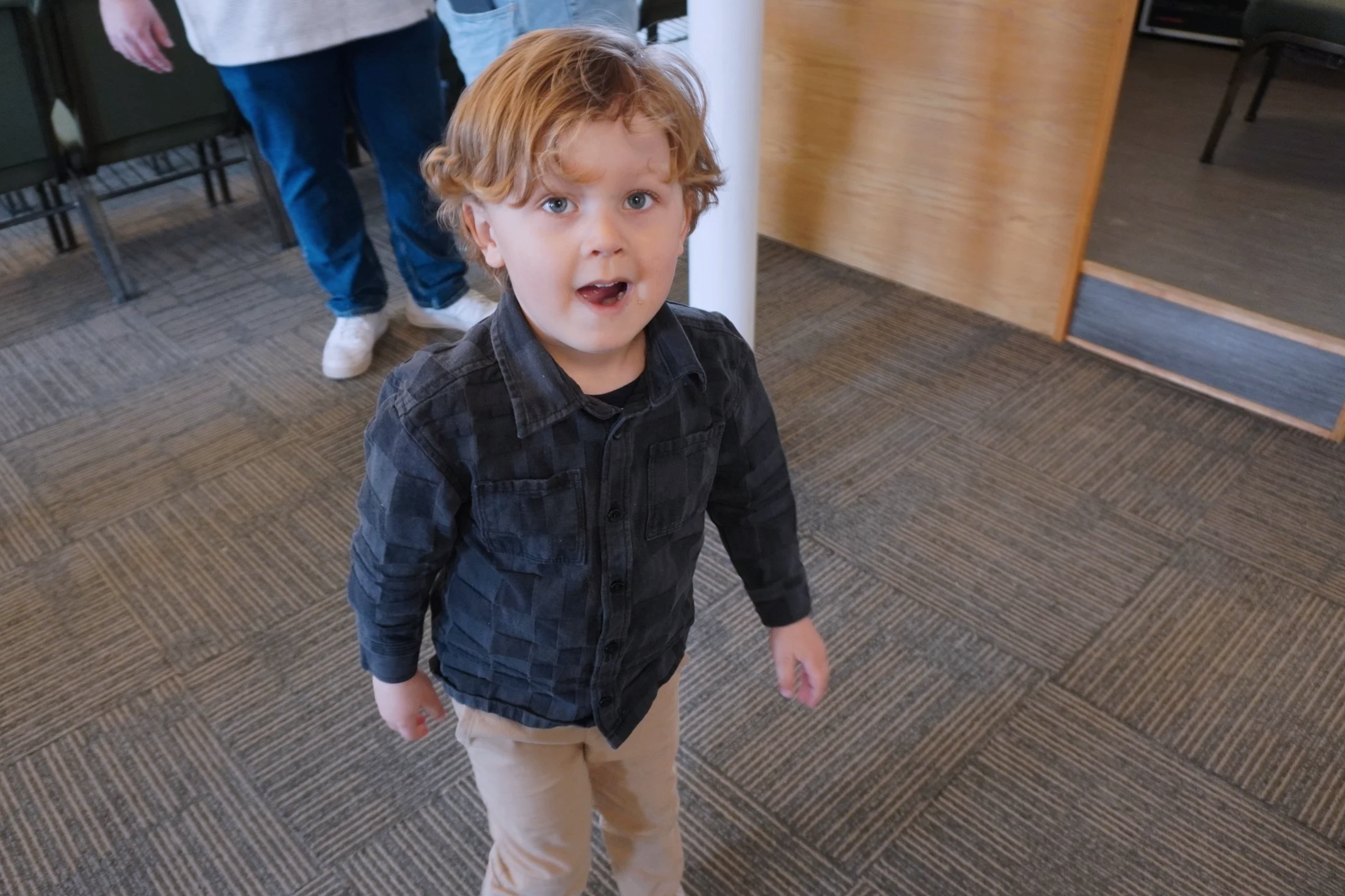 A young boy with red hair and blue eyes looking at the camera, wearing a dark plaid shirt and beige pants, standing indoors on a patterned carpet with a wooden panel and some chairs in the background.
