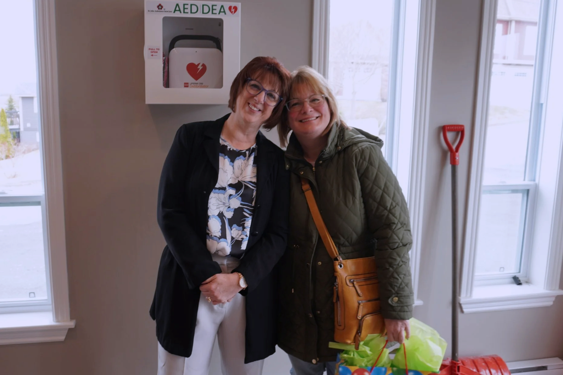 Two women standing inside near a large window, smiling and hugging each other. One woman is dressed in white with a black blazer, and the other is in a green quilted jacket holding shopping bags. There is a fire safety equipment on the wall and a red shovel on the floor.