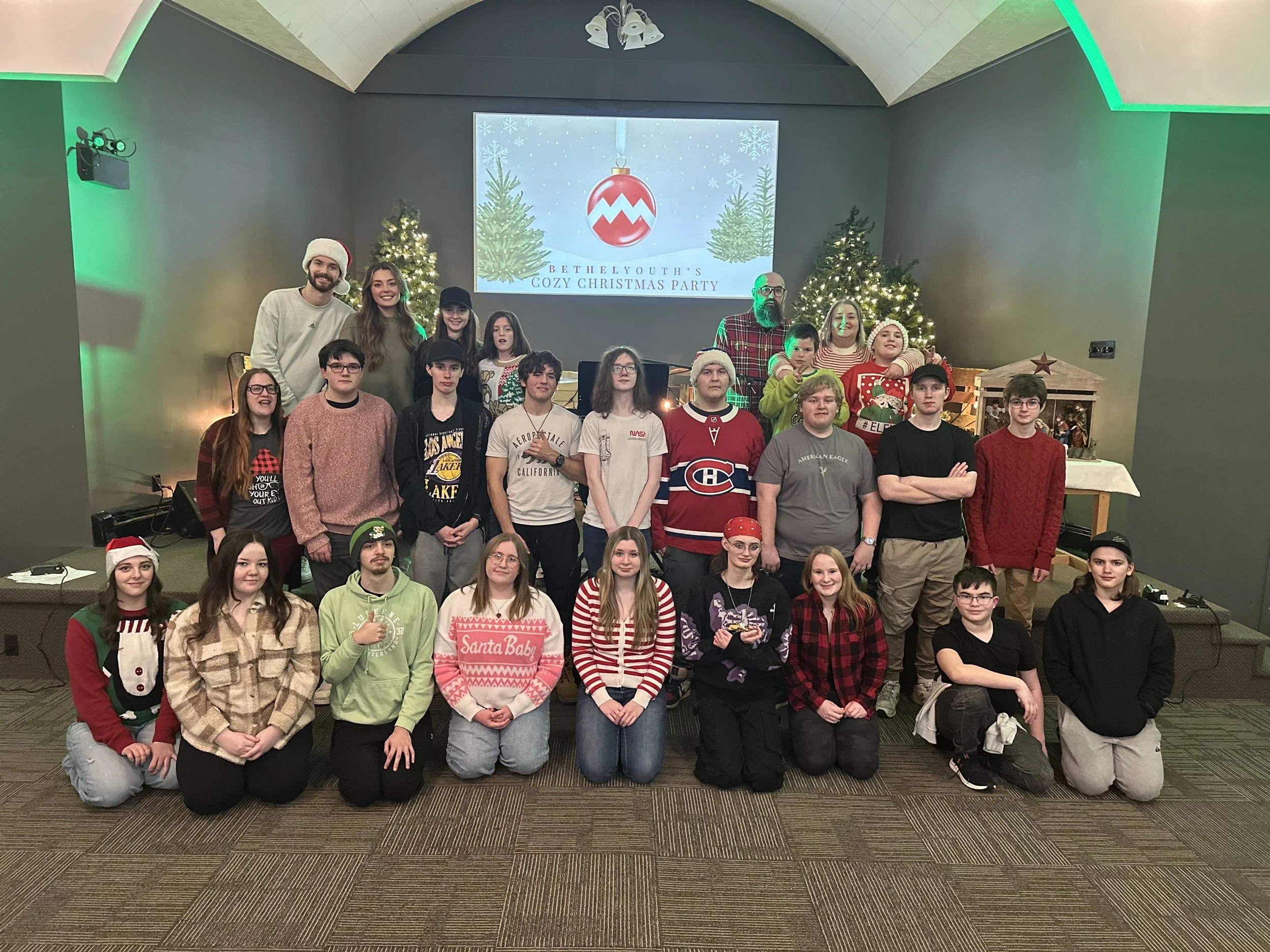 A group of children and adults posing together in a Christmas-themed setting with decorated Christmas trees and festive decorations. A large screen in the background displays a holiday message for a Christmas party.