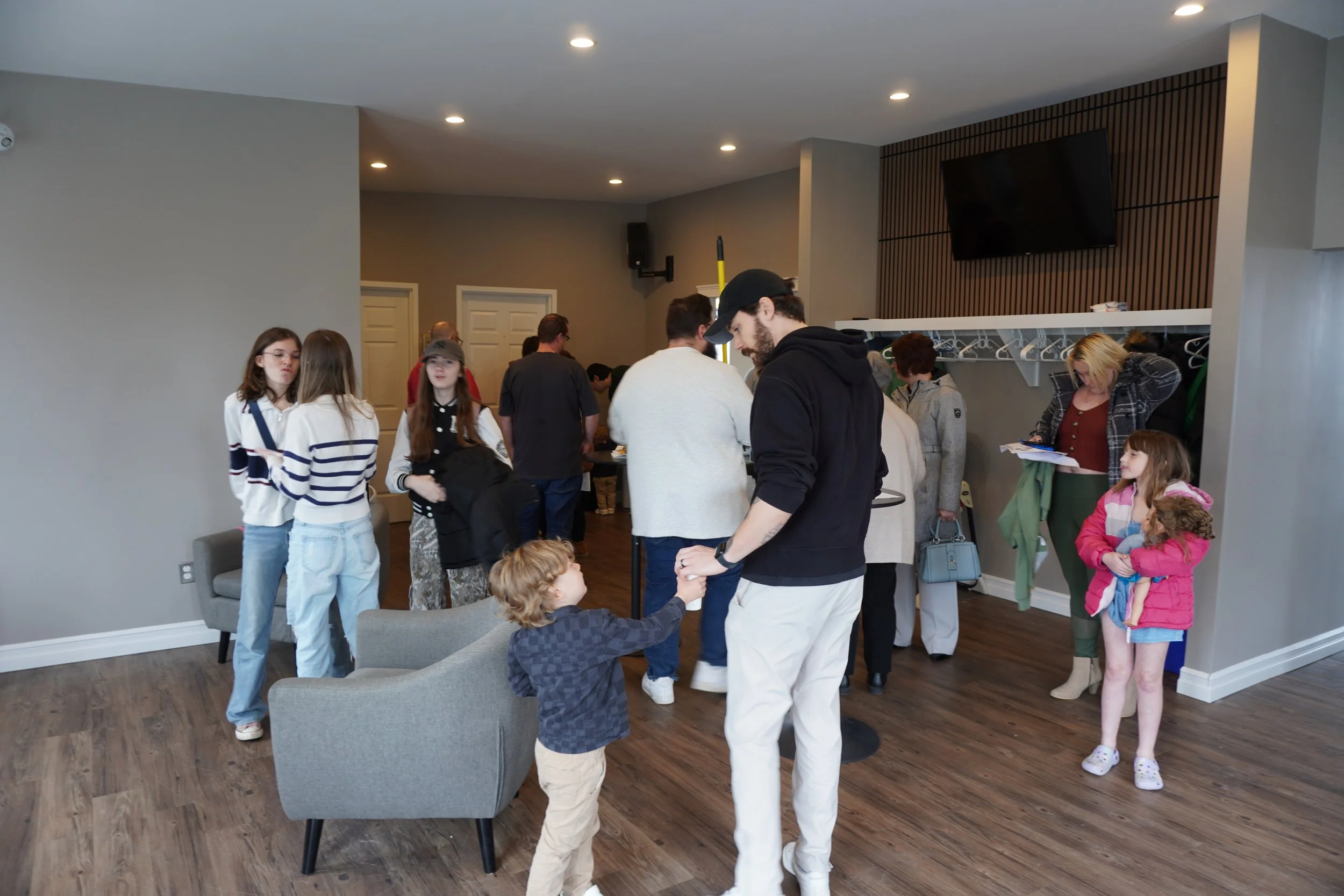People gathering in a modern living room, some standing, some sitting, with children and adults, with a wall-mounted TV and coat hooks along the wall.
