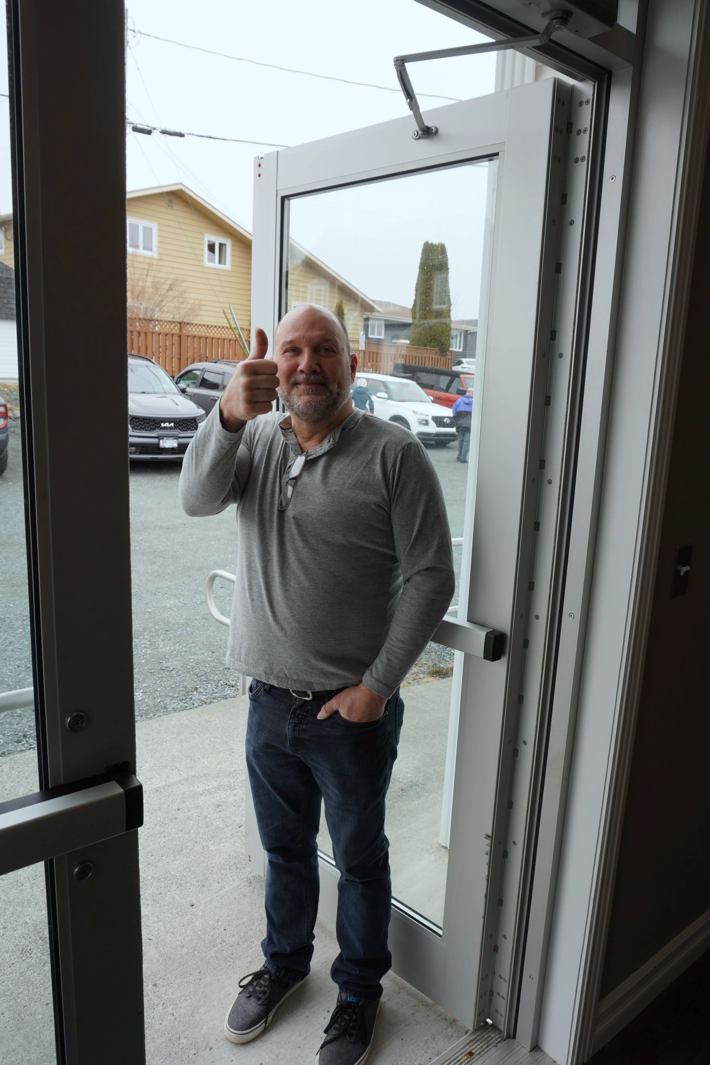 A man standing outside a building entrance, giving a thumbs up, with cars parked in the background and a green tree.