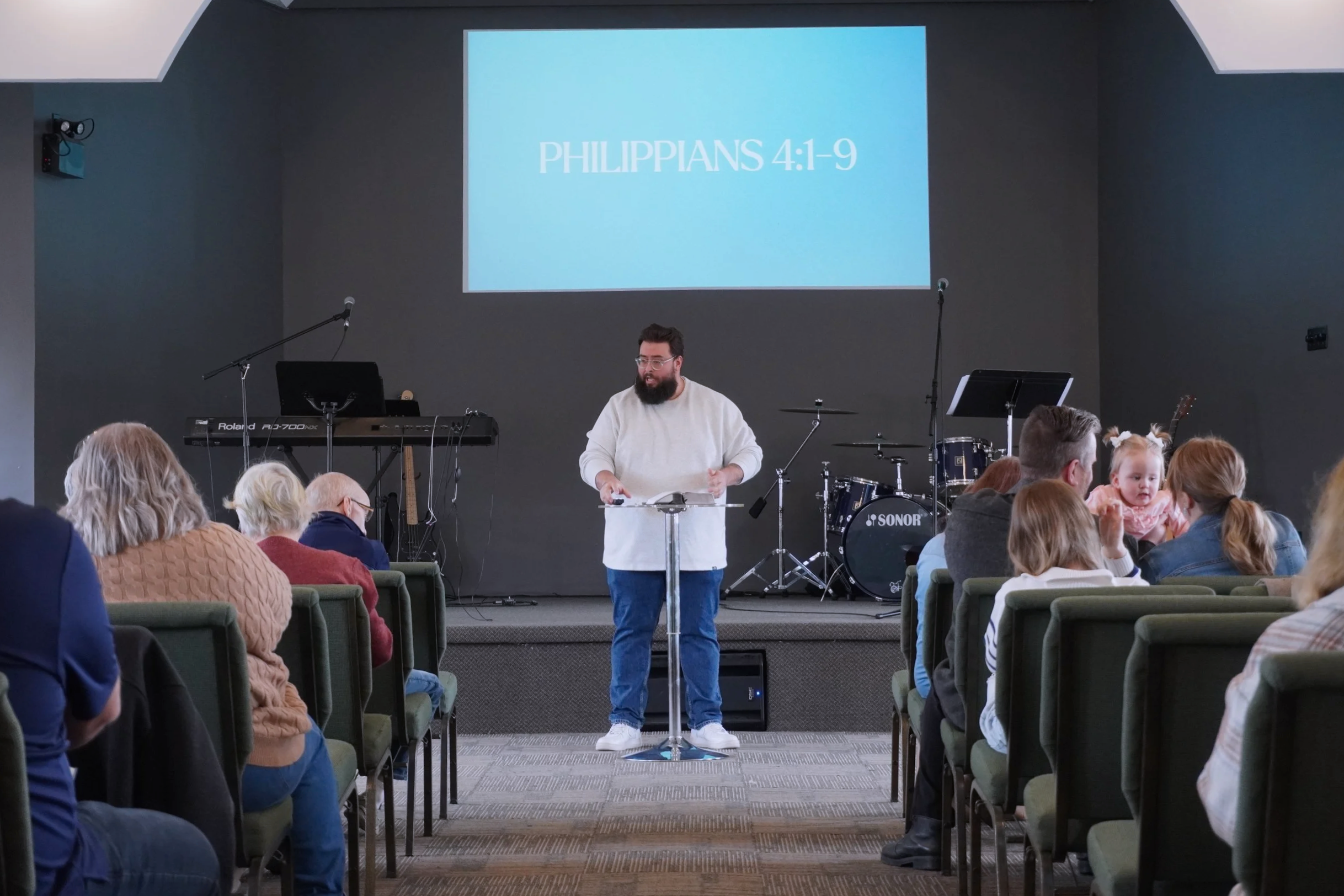 A man with a beard and glasses standing behind a clear lectern, speaking to an audience in a church or presentation setting. The audience is seated facing him, some are children. Behind him, a large screen displays the text 'Philippians 4:1-9'. There is musical equipment, including a keyboard, drums, and microphones, in the background.