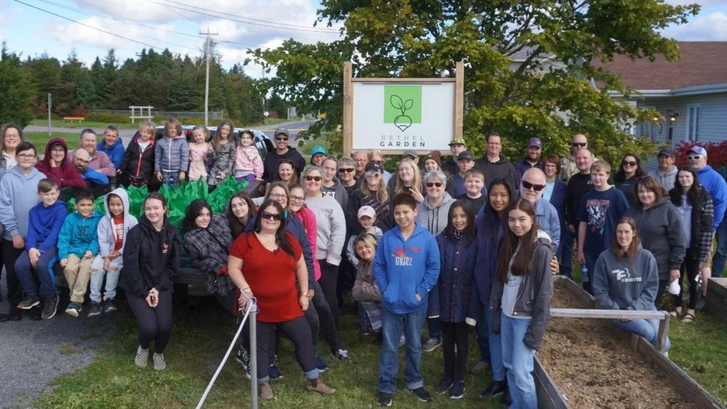 Group of people, including children and adults, gathered in front of a sign that reads 'Bethel Garden' with a logo of a plant sprouting. The group appears to be at a community garden or outdoor event, with trees and houses in the background.