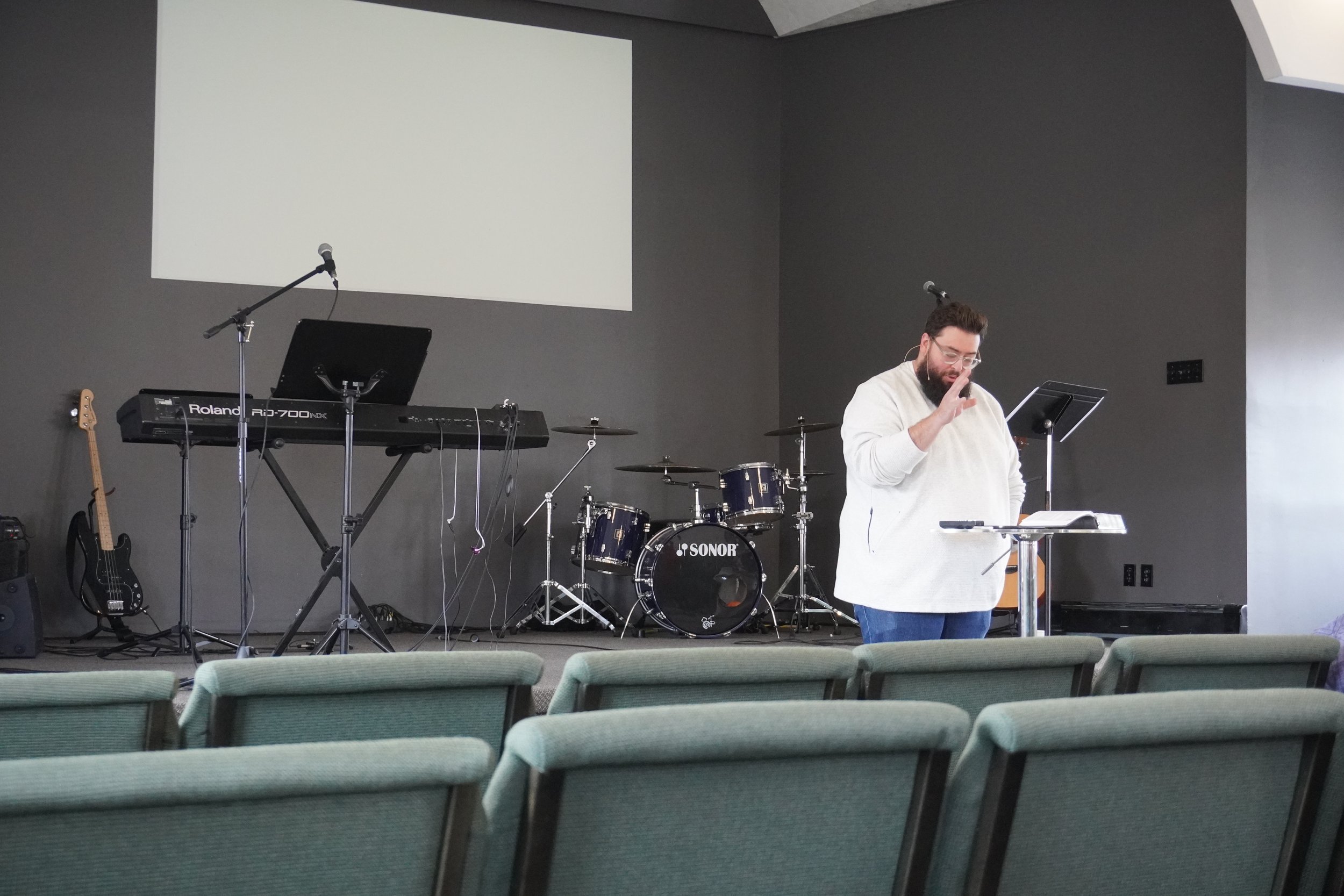 A man with glasses and a beard stands at a music stand on a stage, gesturing with his hand while speaking or singing. Behind him are musical instruments including a drum set, a keyboard, and a guitar. The stage has a large blank projection screen and gray walls.