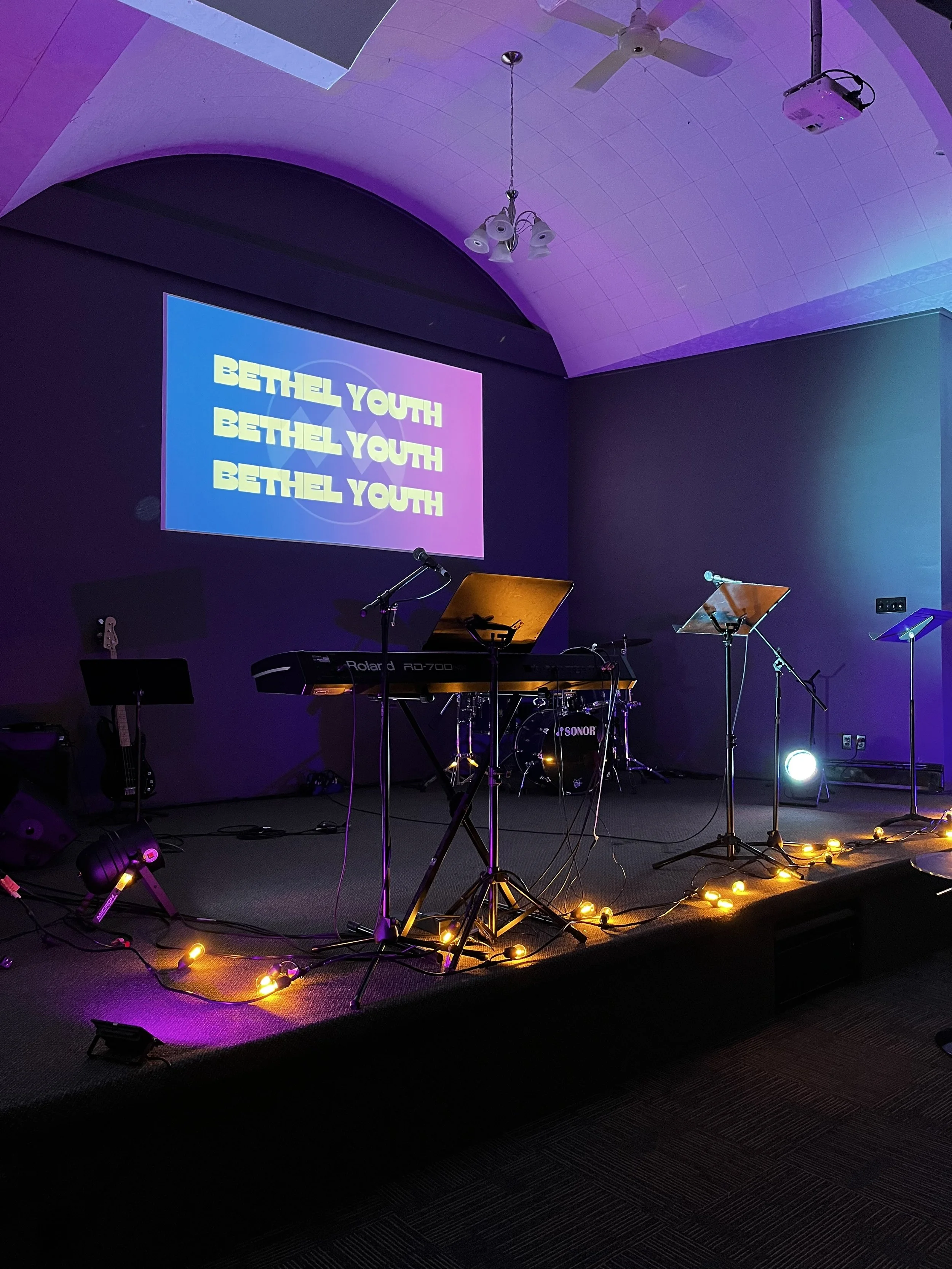 Stage set up with musical instruments and equipment, including a keyboard, microphone, guitars, and music stands. The background features a large screen displaying the words 'Bethel Youth' in bold, colorful letters. The stage is decorated with yellow string lights and is illuminated with purple and blue lighting.