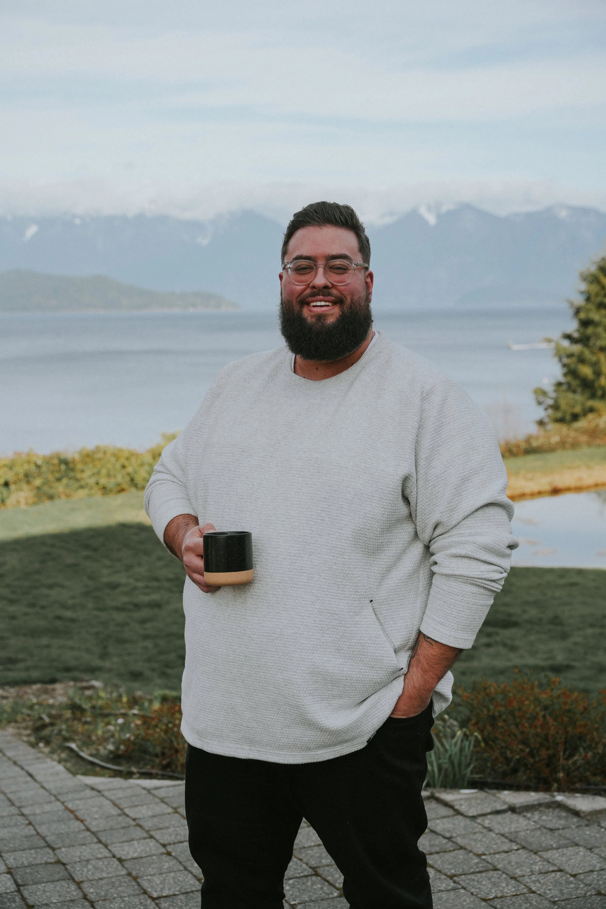 A man with a beard and glasses smiling while holding a cup, standing outdoors with mountains, a lake, and greenery in the background.