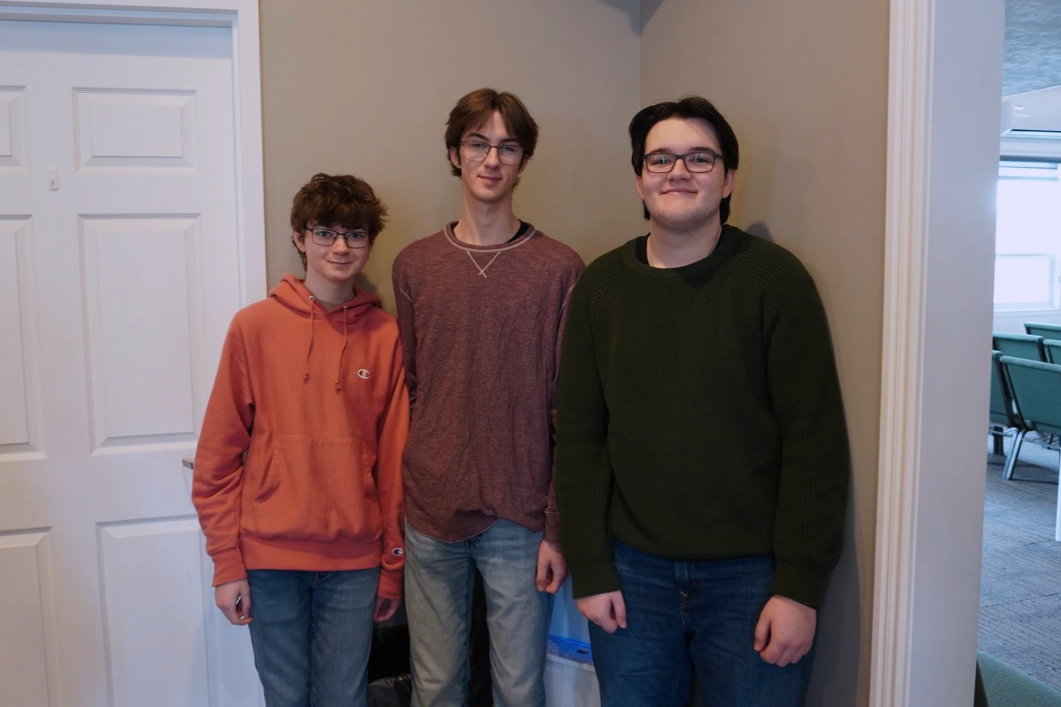 Three males standing together indoors in front of a white door and beige wall. The youngest is on the left wearing an orange hoodie, the middle is in a maroon sweater, and the oldest on the right wears a dark green sweater and glasses.