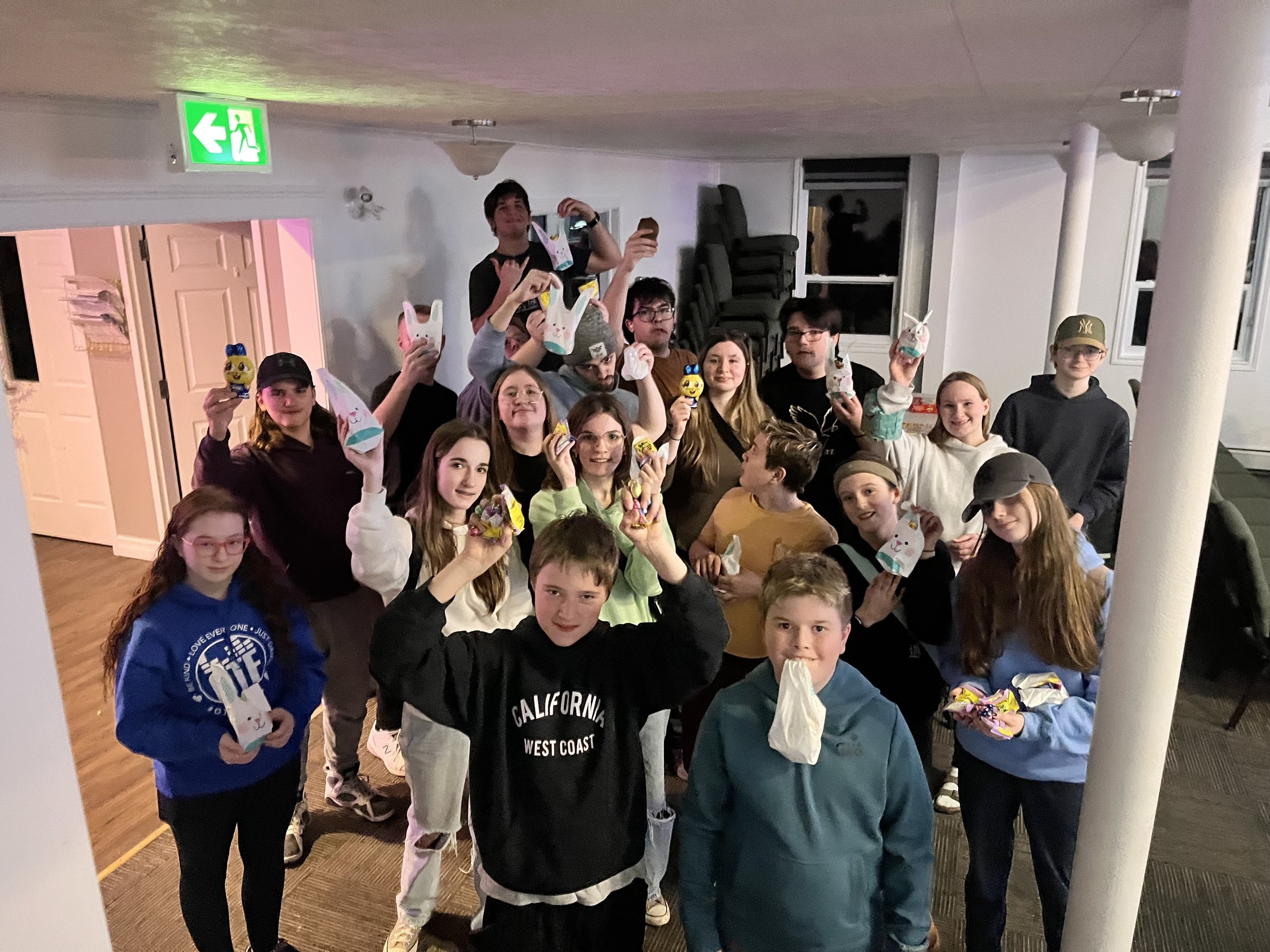 A group of children and teenagers, smiling and holding decorated paper bunny crafts, gathered indoors during a festive celebration.