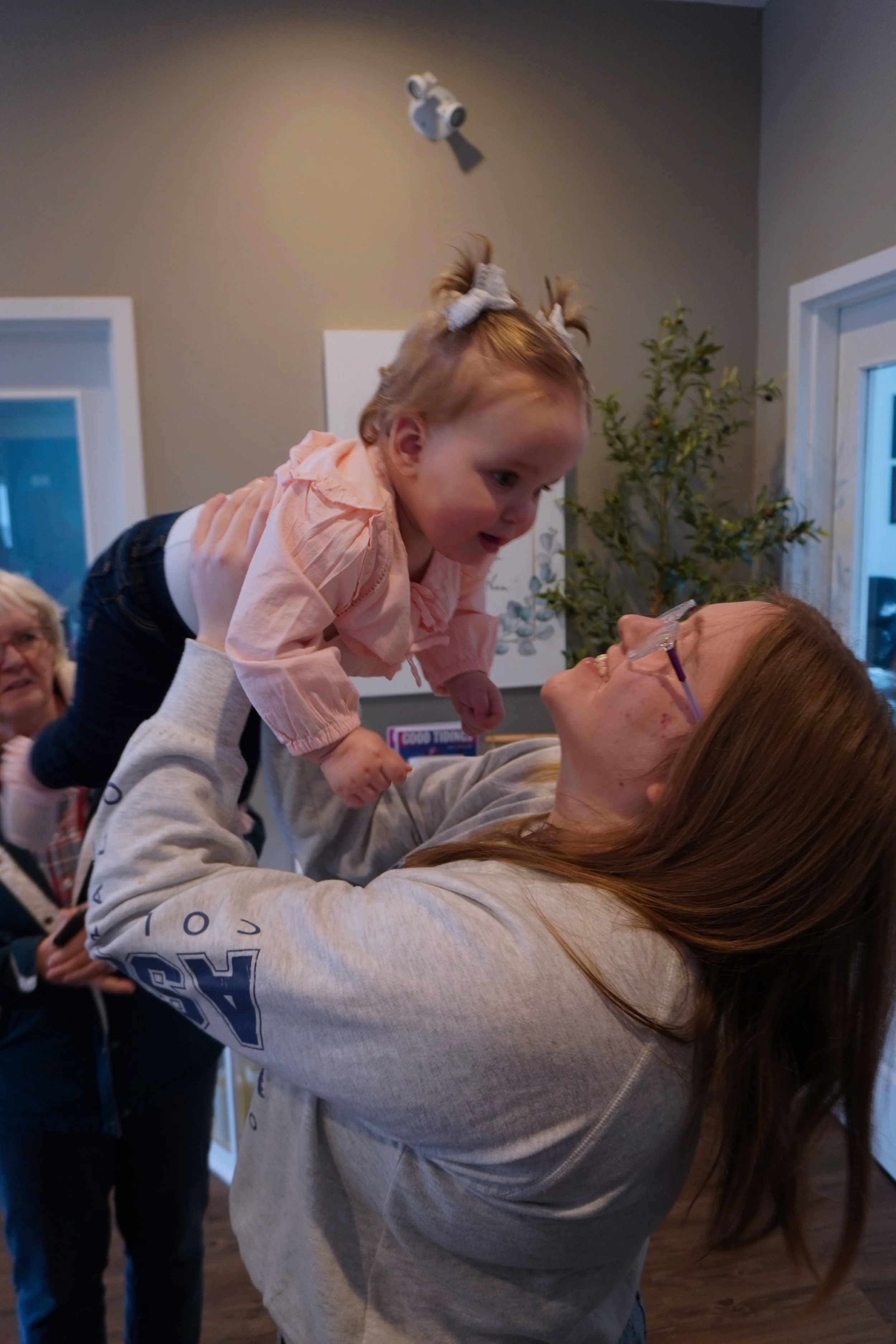 A young girl with bows in her hair is being held up on an older woman's shoulders inside a room. The girl is looking down at the woman, and they both appear happy. In the background, there are other people and indoor decorations.