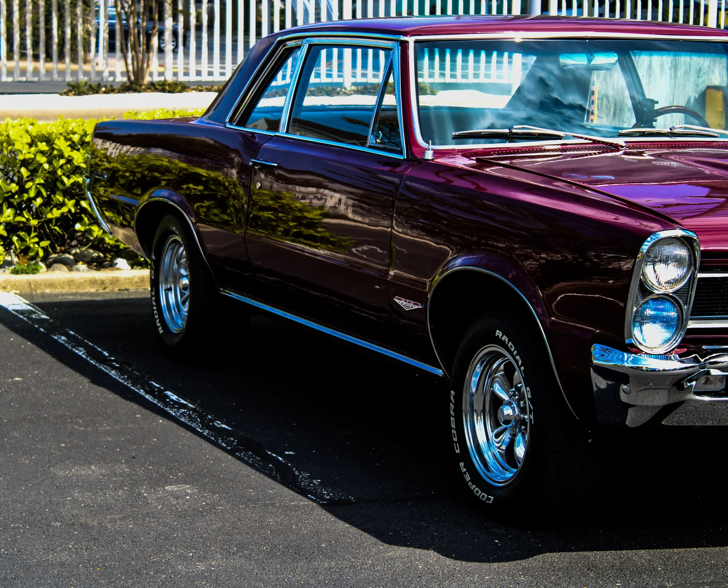 A vintage maroon and black two-tone station wagon car parked in a parking lot, with a white fence and some trees in the background.