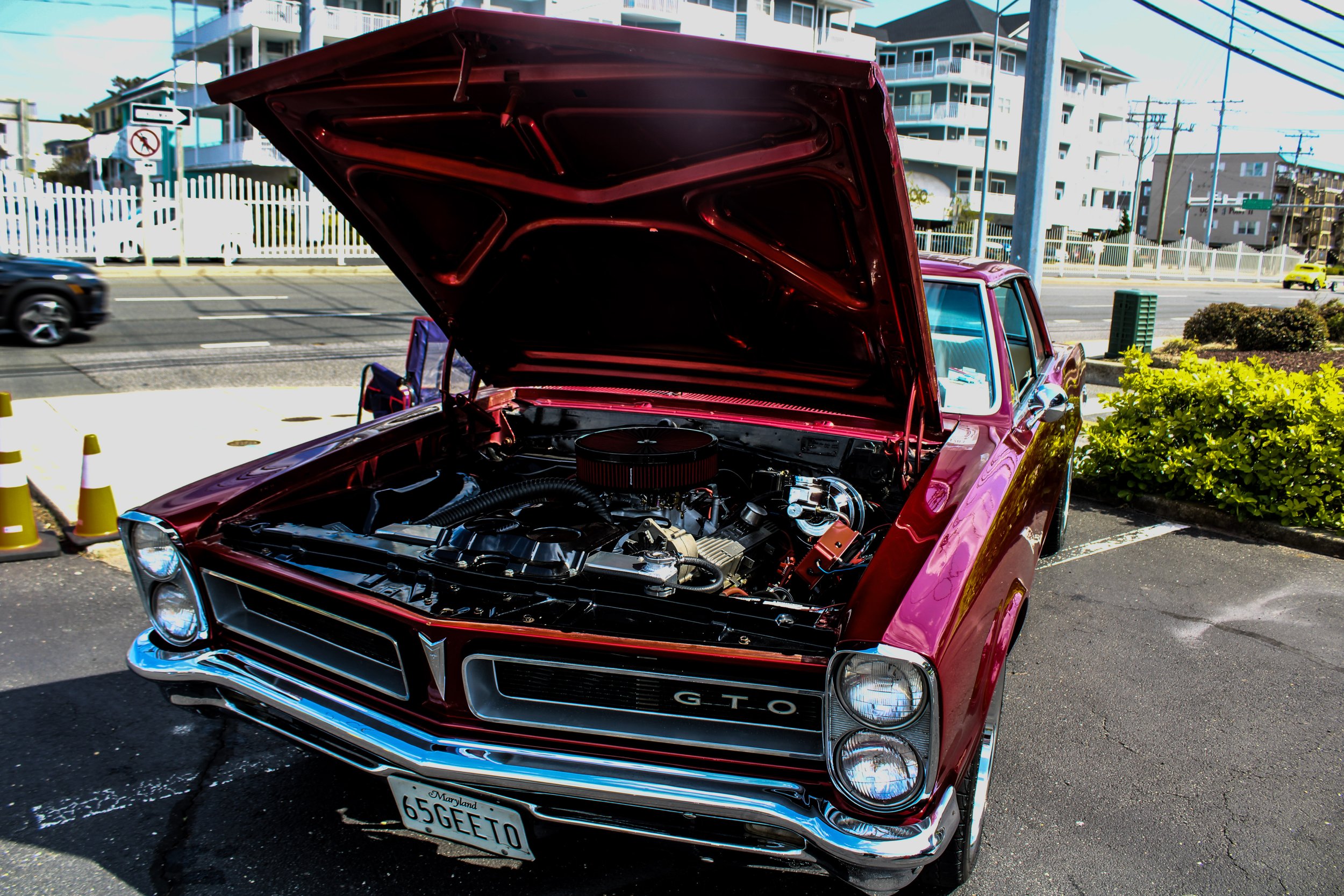 A red classic muscle car with its hood open, showing the engine, parked on a city street near modern apartment buildings.